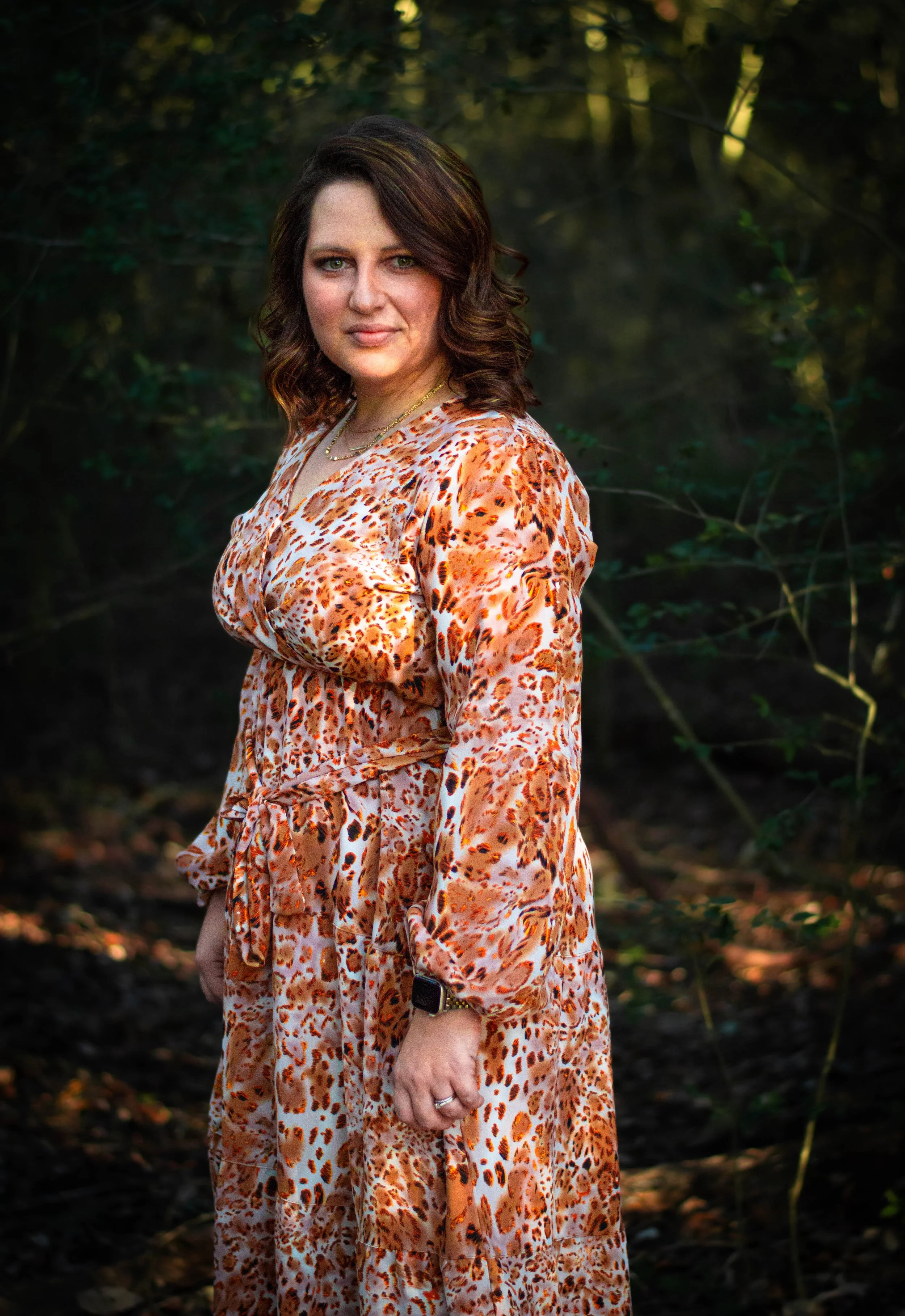 A woman with shoulder-length brown hair and light skin standing outdoors in a wooded area, wearing a long dress with a leopard print pattern in shades of orange, brown, and cream. She is looking at the camera with a slight smile, with sunlight filter