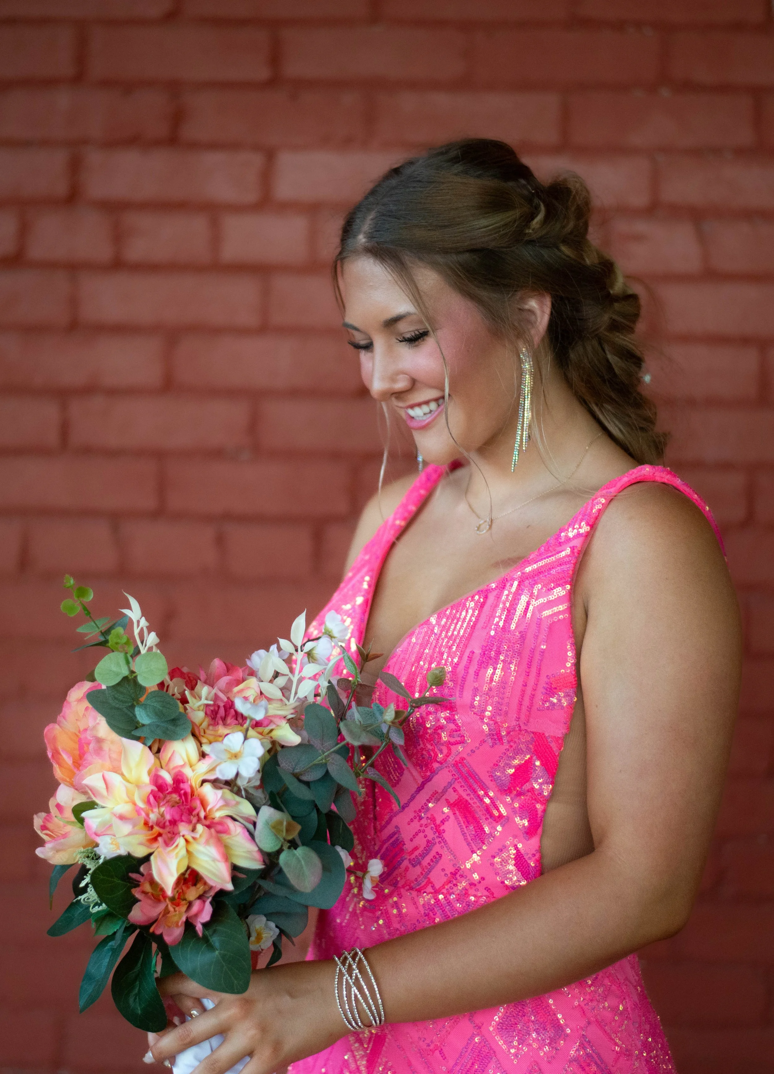 A woman in a bright pink, sequined dress holding a bouquet of pink and white flowers, standing in front of a red brick wall.