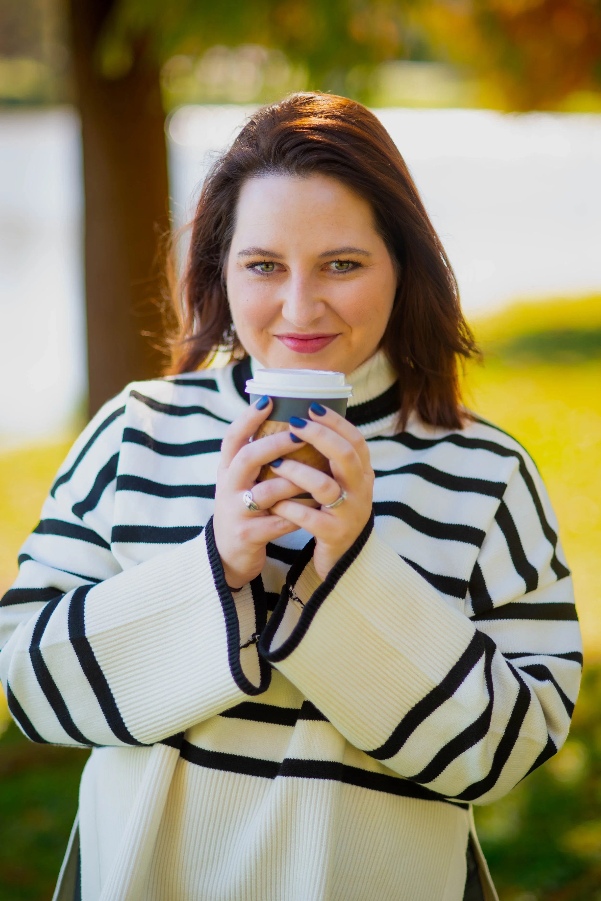 A woman with shoulder-length brown hair and blue eyes, wearing a white and black striped sweater, is holding a disposable coffee cup with both hands outdoors near a lake or river, with trees and green grass in the background.
