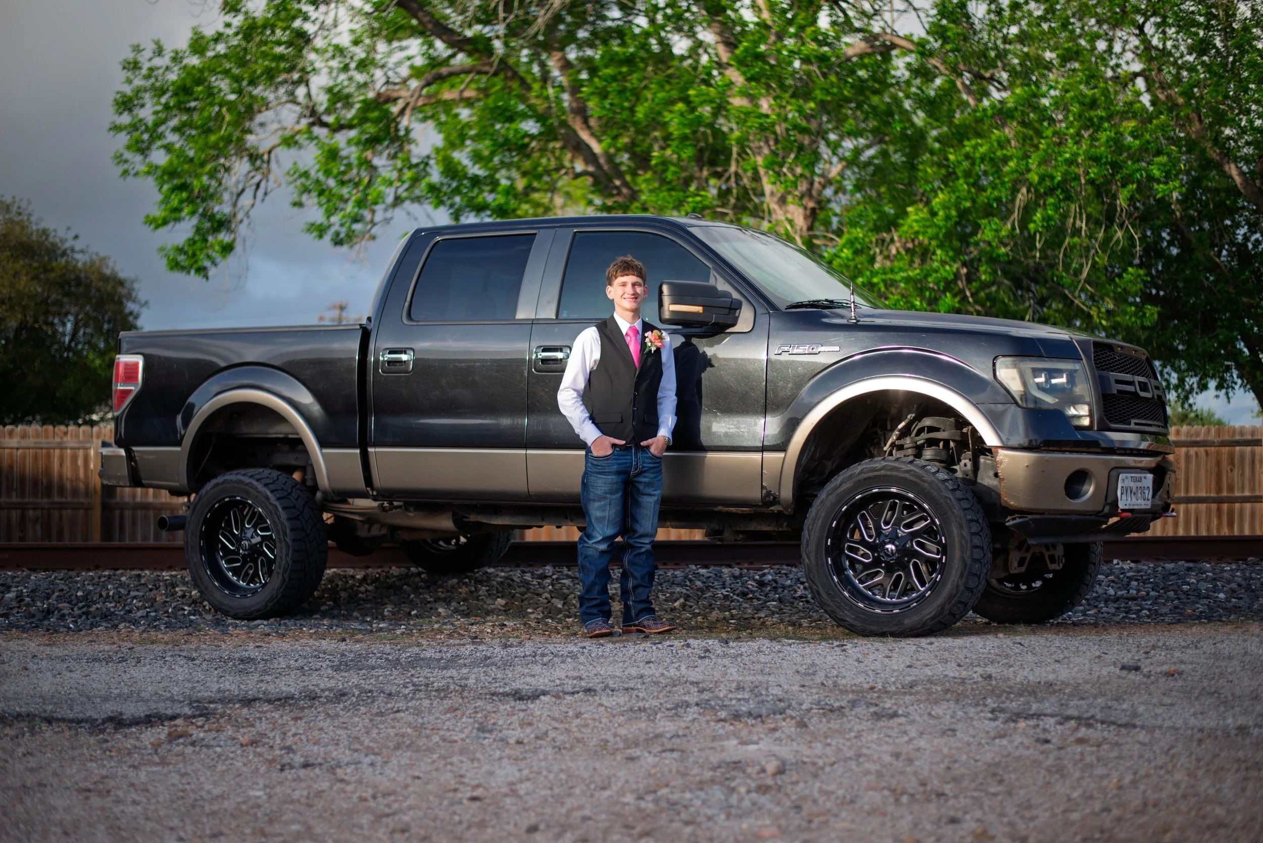 Young man in formal attire standing next to a lifted black pickup truck outdoors near trees and a wooden fence.
