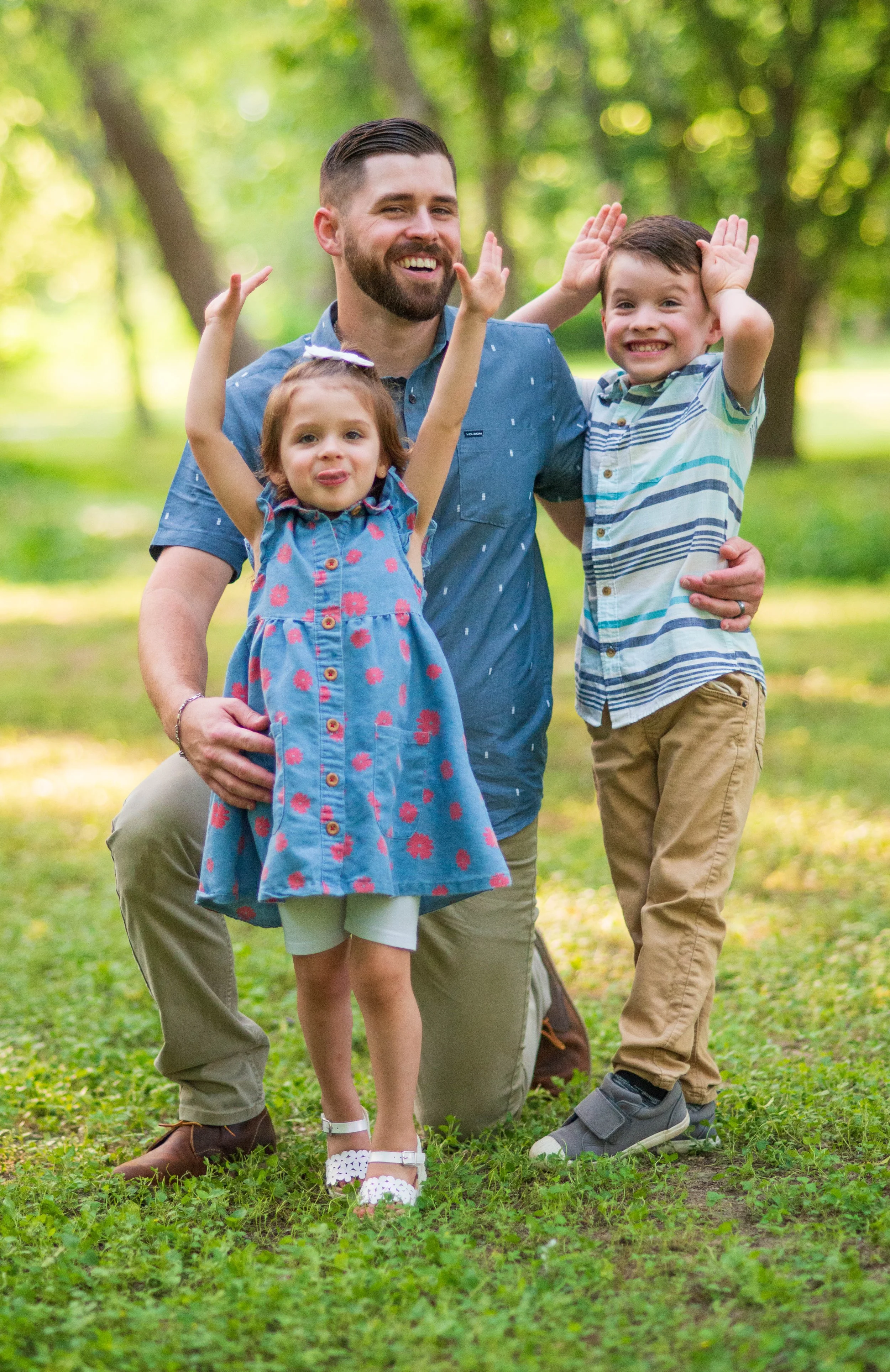 A happy man with a beard kneeling in a park with a young girl and boy, all smiling and making playful gestures with their hands, surrounded by green trees and grass.