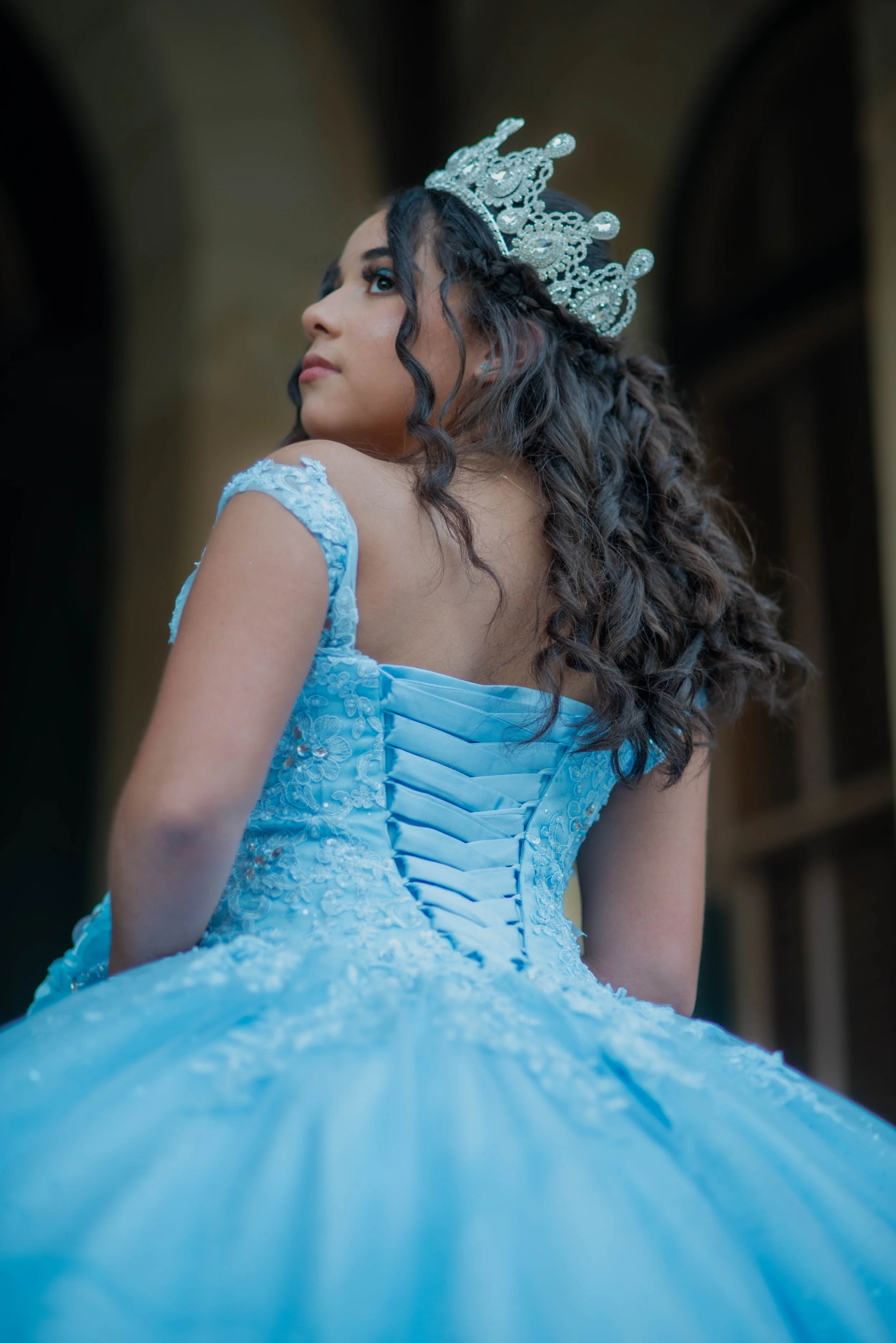 A young woman in a blue ball gown with lace details and a lace-up back, wearing a silver tiara, looking over her shoulder with dark curly hair.