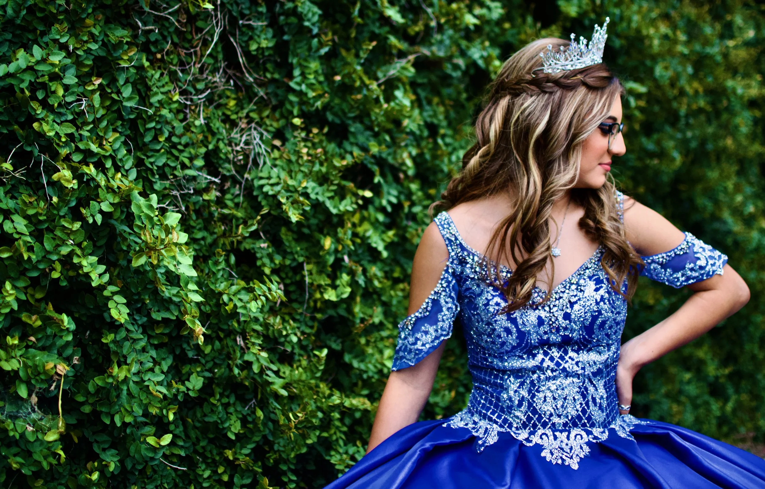 Young woman in a blue, beaded, off-the-shoulder gown with a crown and necklace, standing sideways against a green leafy background.