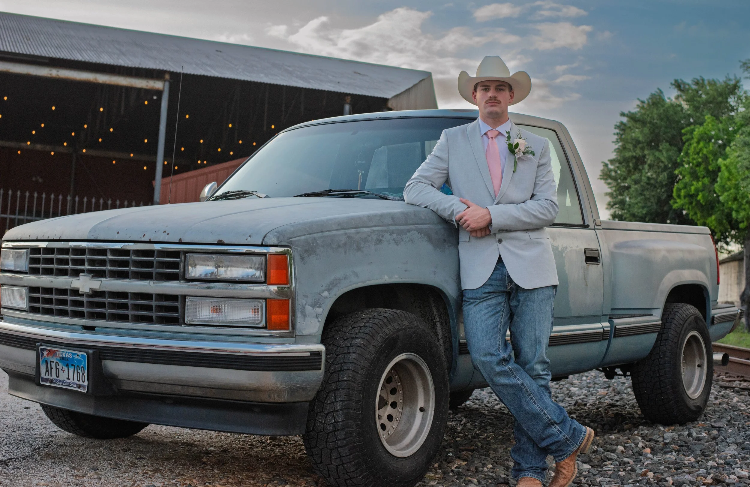 A man in a light gray suit and cowboy hat leaning against an old, weathered pickup truck with a Texas license plate, outdoors in the evening.