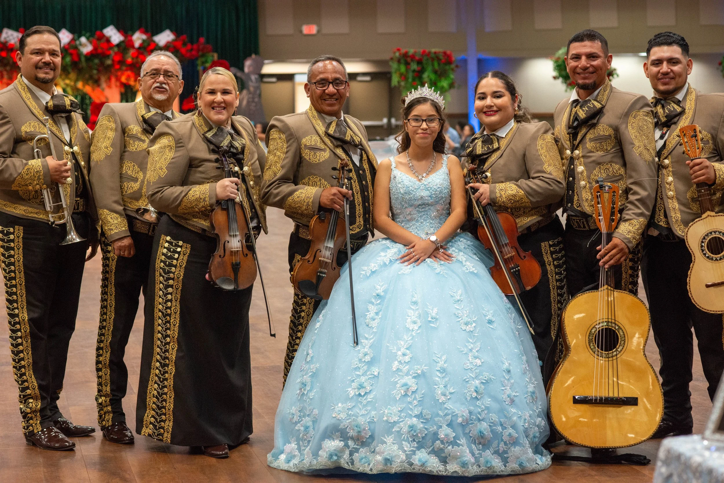 A young woman in a light blue, floral-adorned dress, wearing a tiara, is sitting among several musicians in traditional Mexican mariachi outfits. The musicians are holding various instruments like violins, guitars, and trumpets, in a festive indoor s