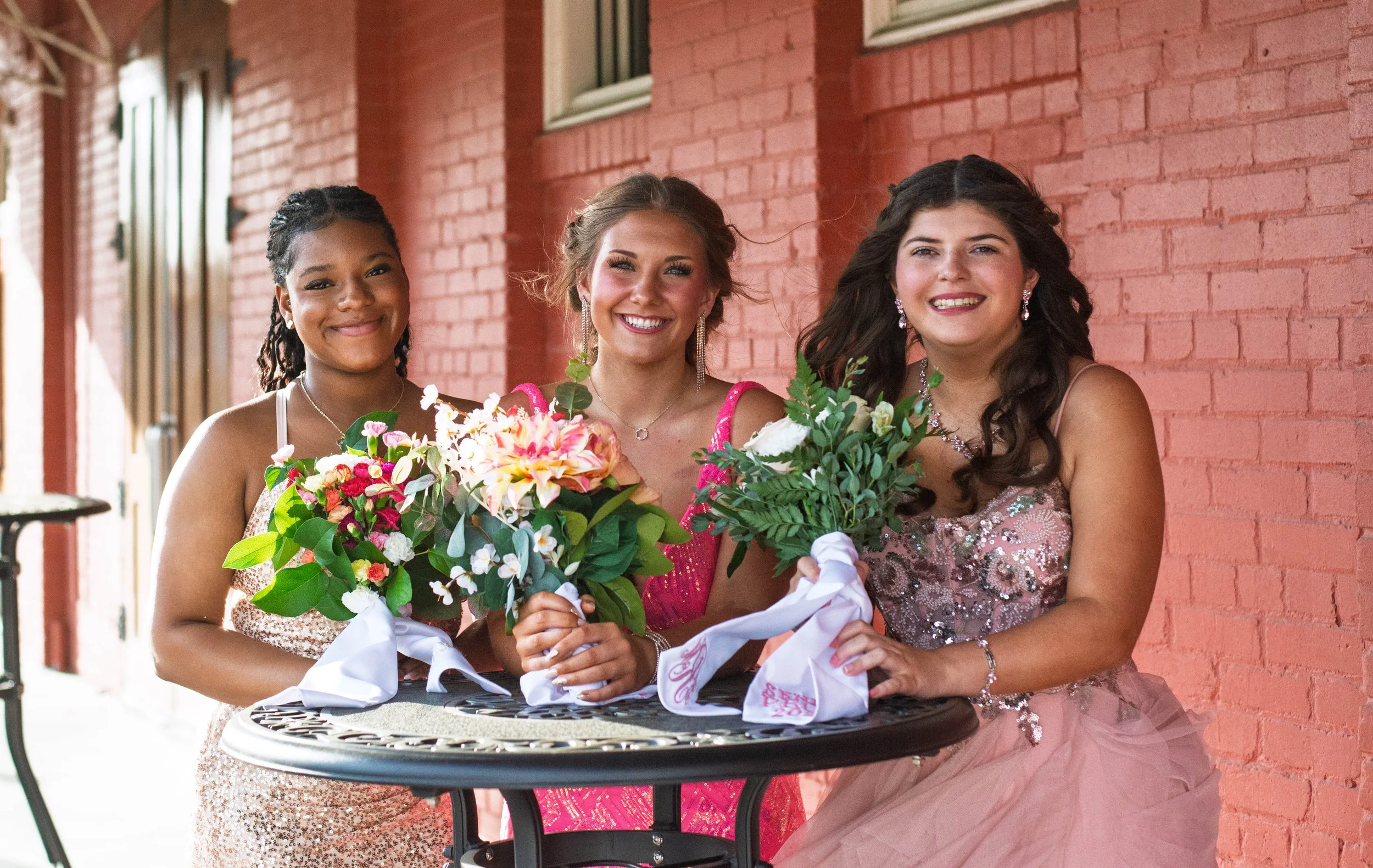 Three women in formal dresses smiling and holding bouquets of flowers, standing behind a round table against a red brick wall.
