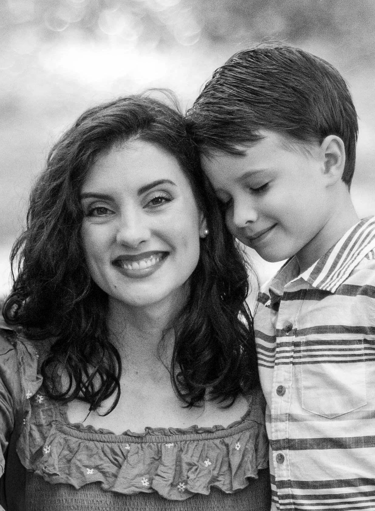 A woman with dark wavy hair smiling, standing next to a boy with closed eyes and short dark hair, both outdoors.