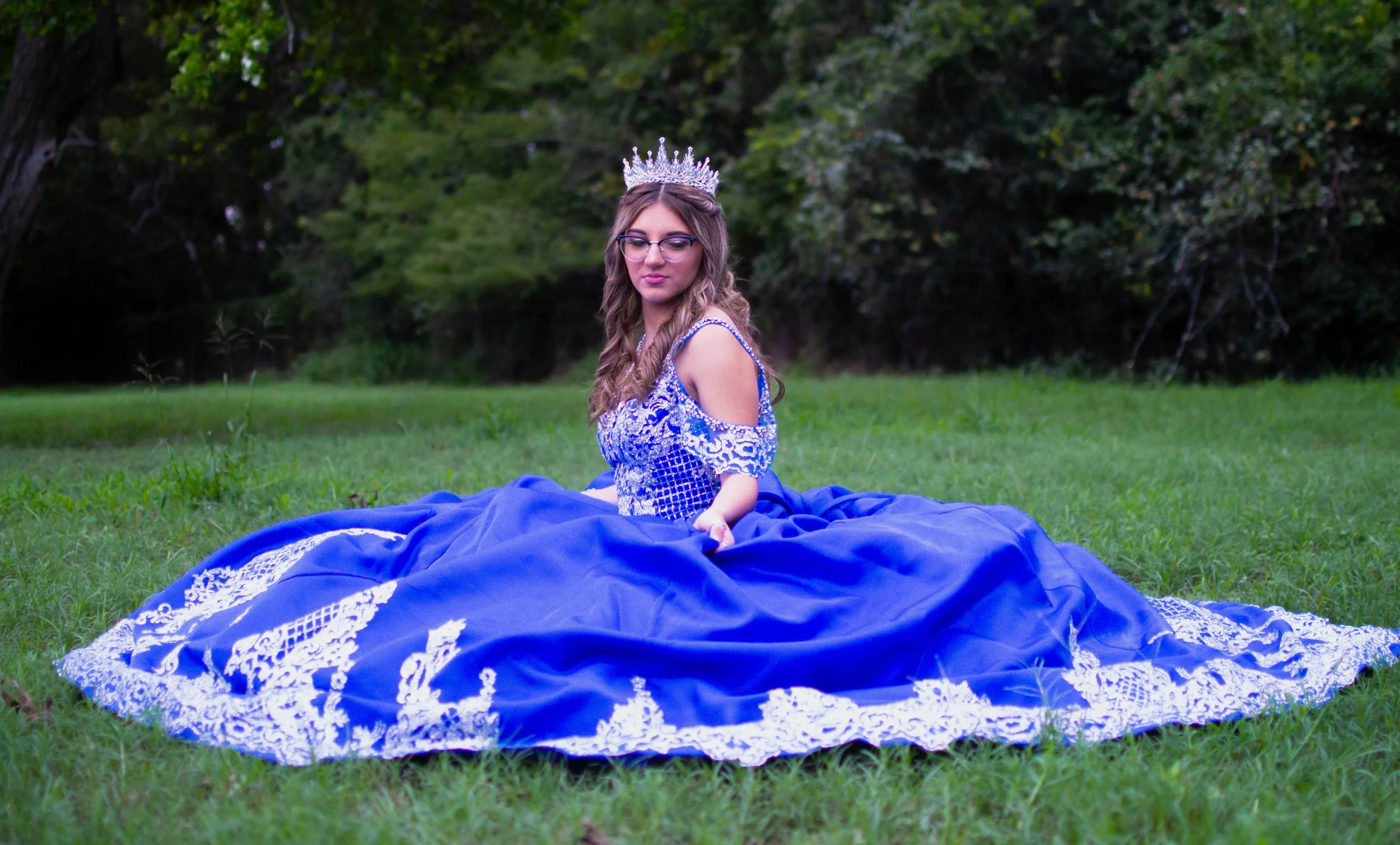 Young woman sitting on grass in a blue dress with lace details, wearing a crown, outdoors in a green park or forest.