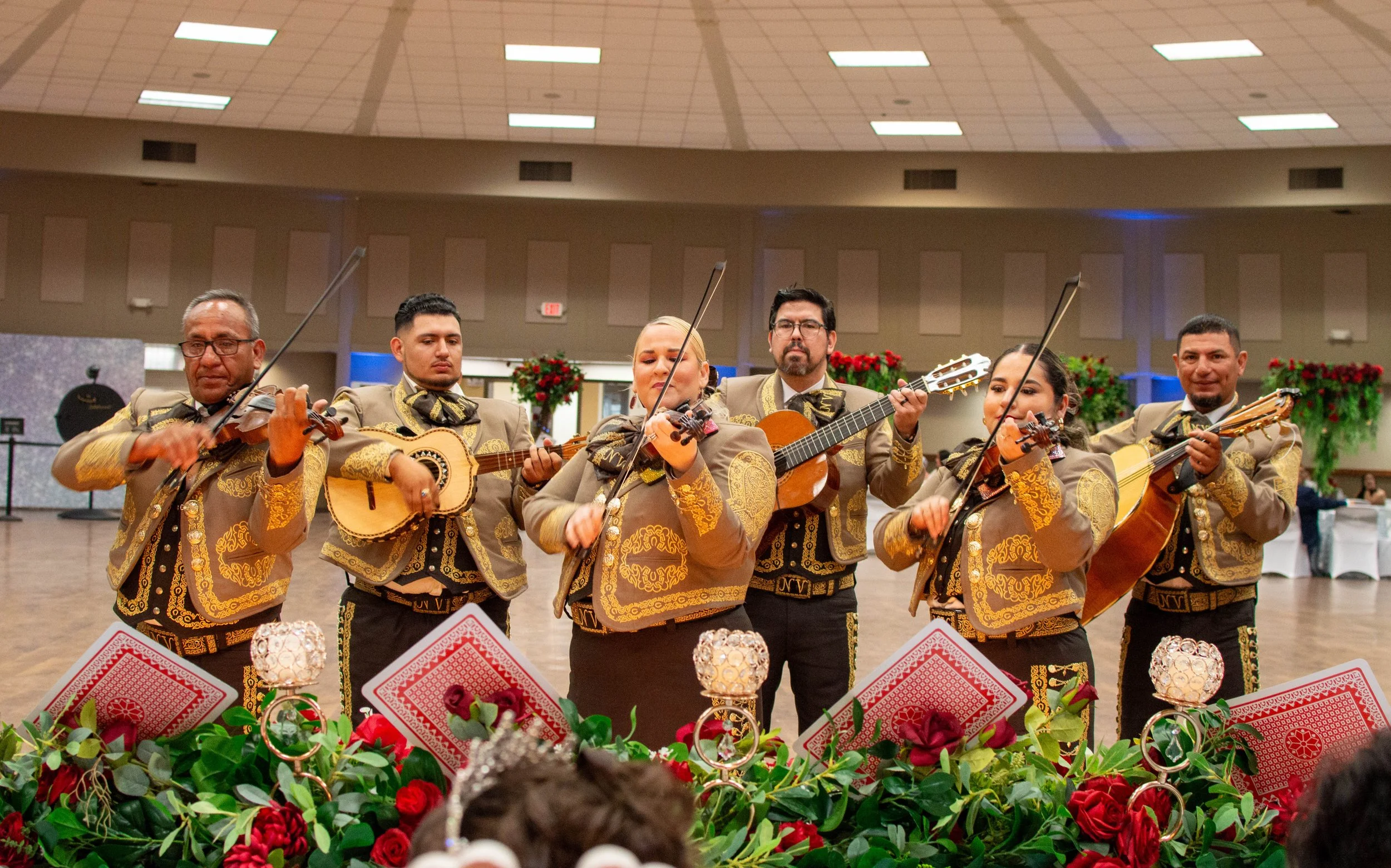 A group of six musicians dressed in traditional Mexican charro outfits playing guitars and violins at a festive event with floral decorations and a decorated table front.