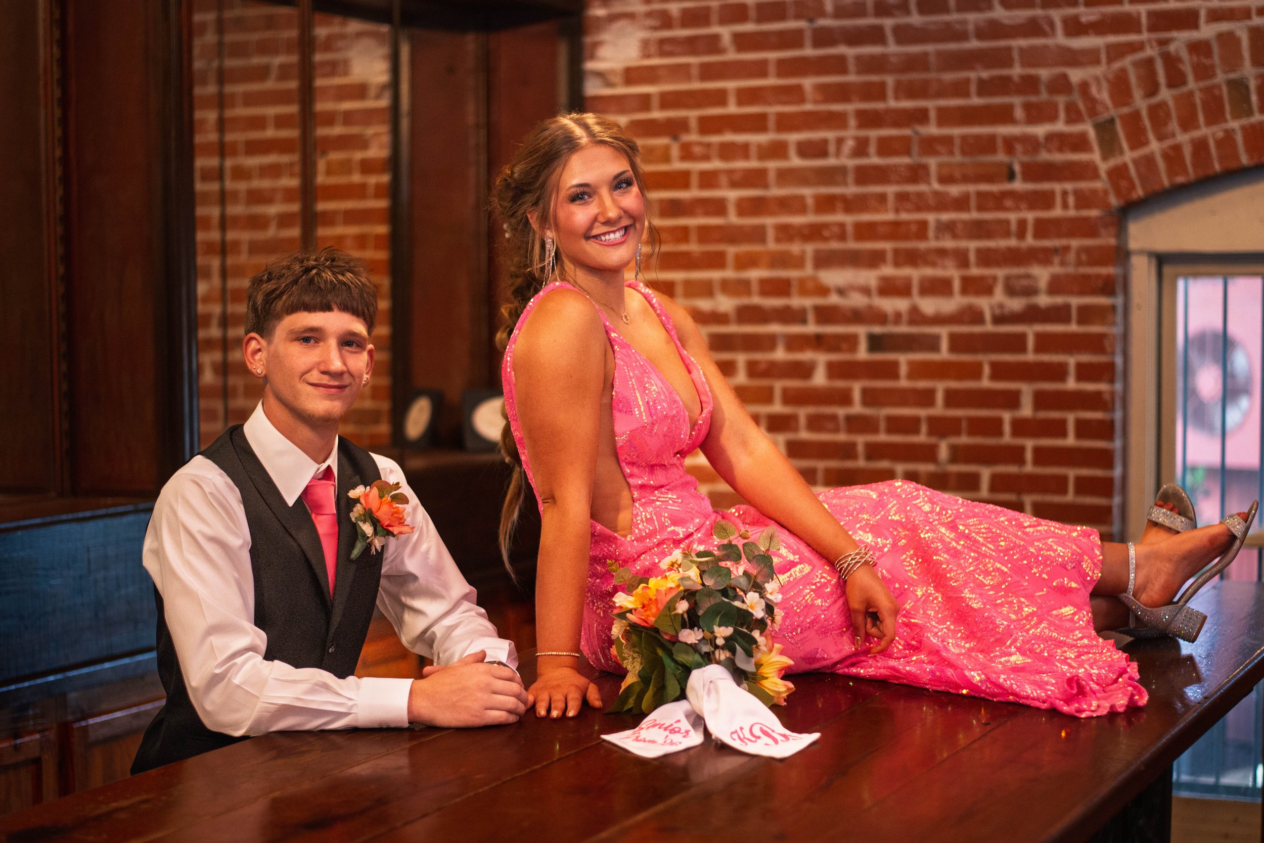 A young man and woman in formal attire sit on a wooden table in a brick-walled room. The woman is wearing a pink dress with shiny details, silver high heels, and jewelry. The young man is wearing a black vest, white shirt, and a pink tie, with a bout