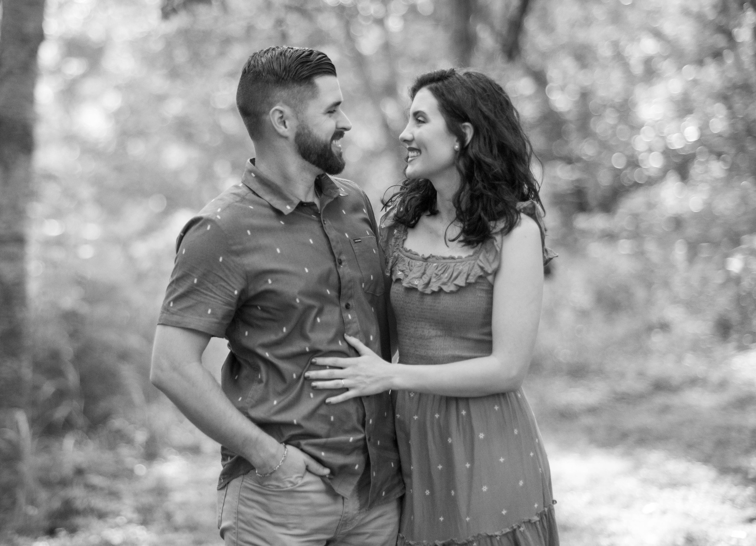 A black and white photograph of a smiling couple standing close to each other outdoors, looking into each other's eyes. The man has a beard and is wearing a short-sleeved button-up shirt, while the woman has dark, wavy hair and is wearing a sleeveles