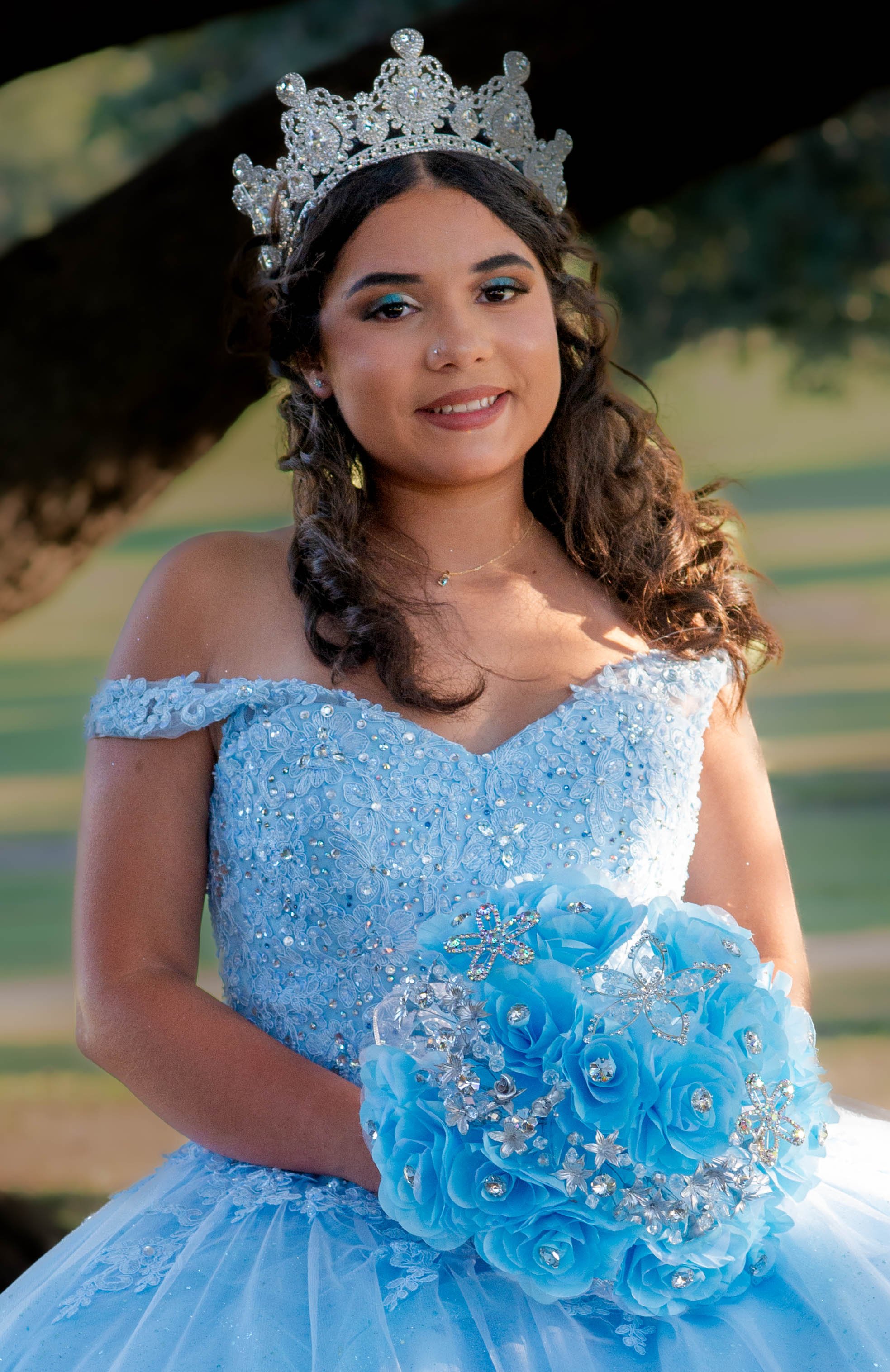 Young woman dressed in a light blue wedding gown, wearing a silver tiara, holding a bouquet of blue fabric roses decorated with rhinestones and floral embellishments, smiling outdoors with a blurred green background.
