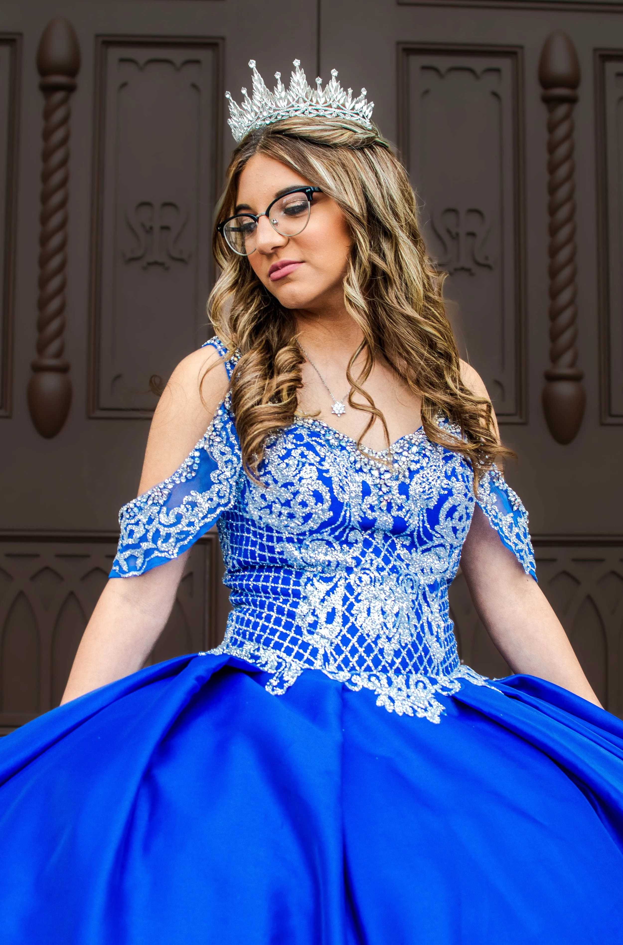 Young woman in a blue gown with silver embroidery, wearing a tiara and glasses, standing in front of a dark wooden background.