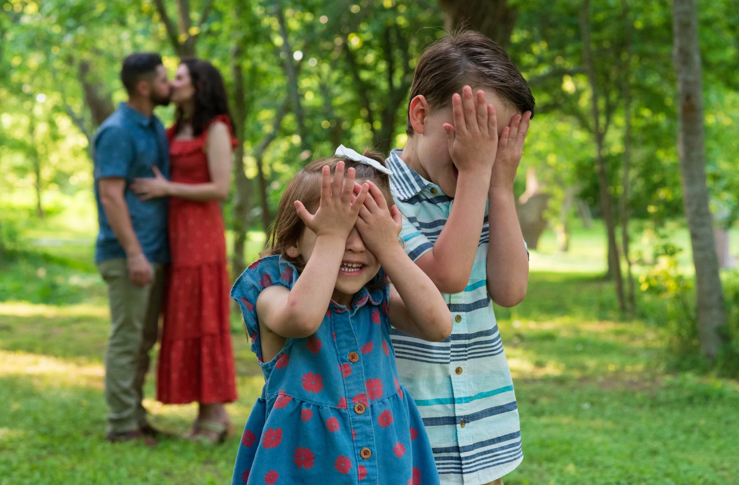 Two children, a girl in a blue dress with red flowers and a boy in a striped shirt, cover their eyes and smile. In the blurred background, a couple is kissing in a park with green trees.