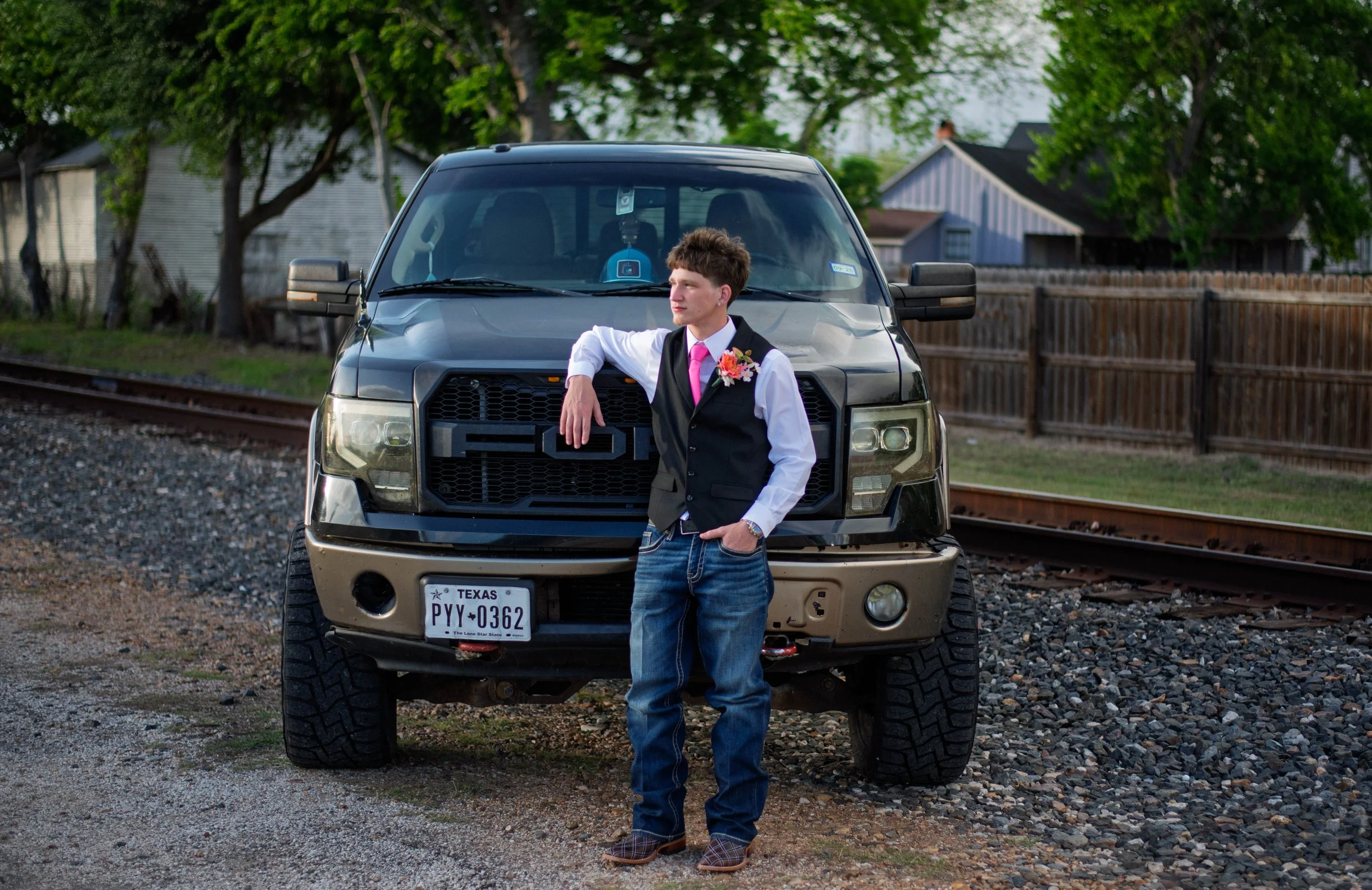 A young man in a formal vest and pink tie leaning on the front of a black pickup truck on a gravel area beside railroad tracks and wooden fences, with trees and houses in the background.