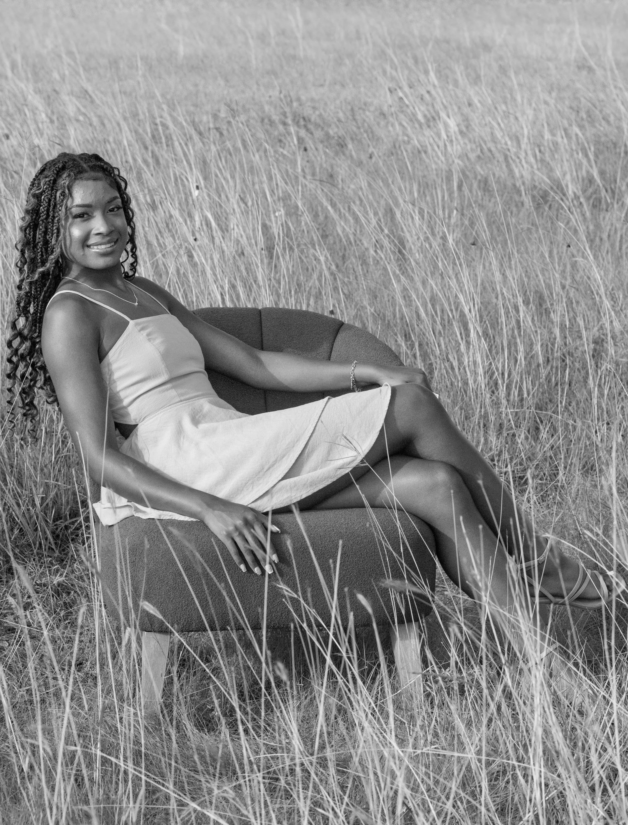 A woman sitting on a cushioned chair in a field of tall grass, smiling, wearing a light-colored dress with spaghetti straps.