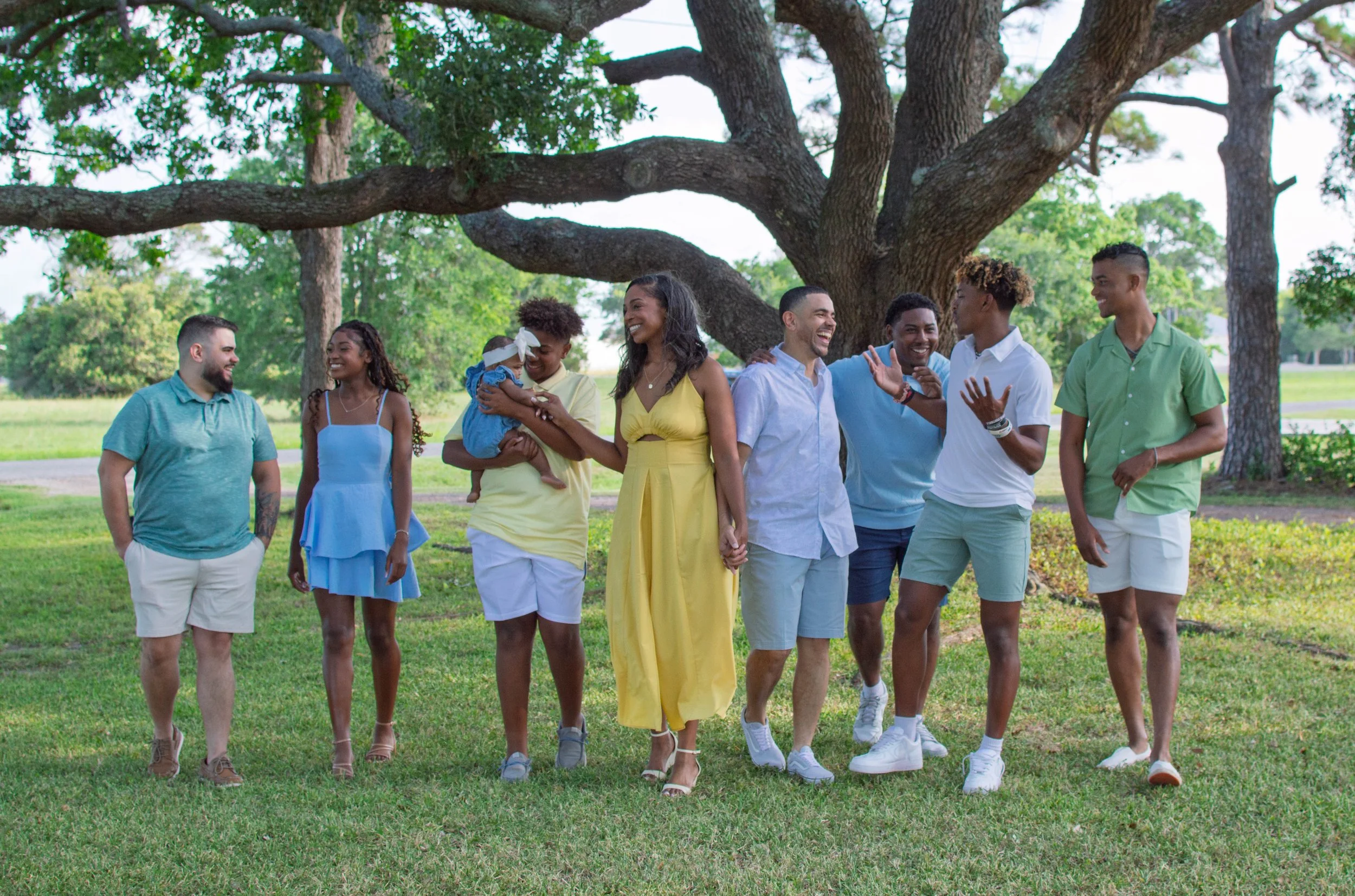 Group of nine diverse young adults and one child walking and laughing together outdoors under a large tree