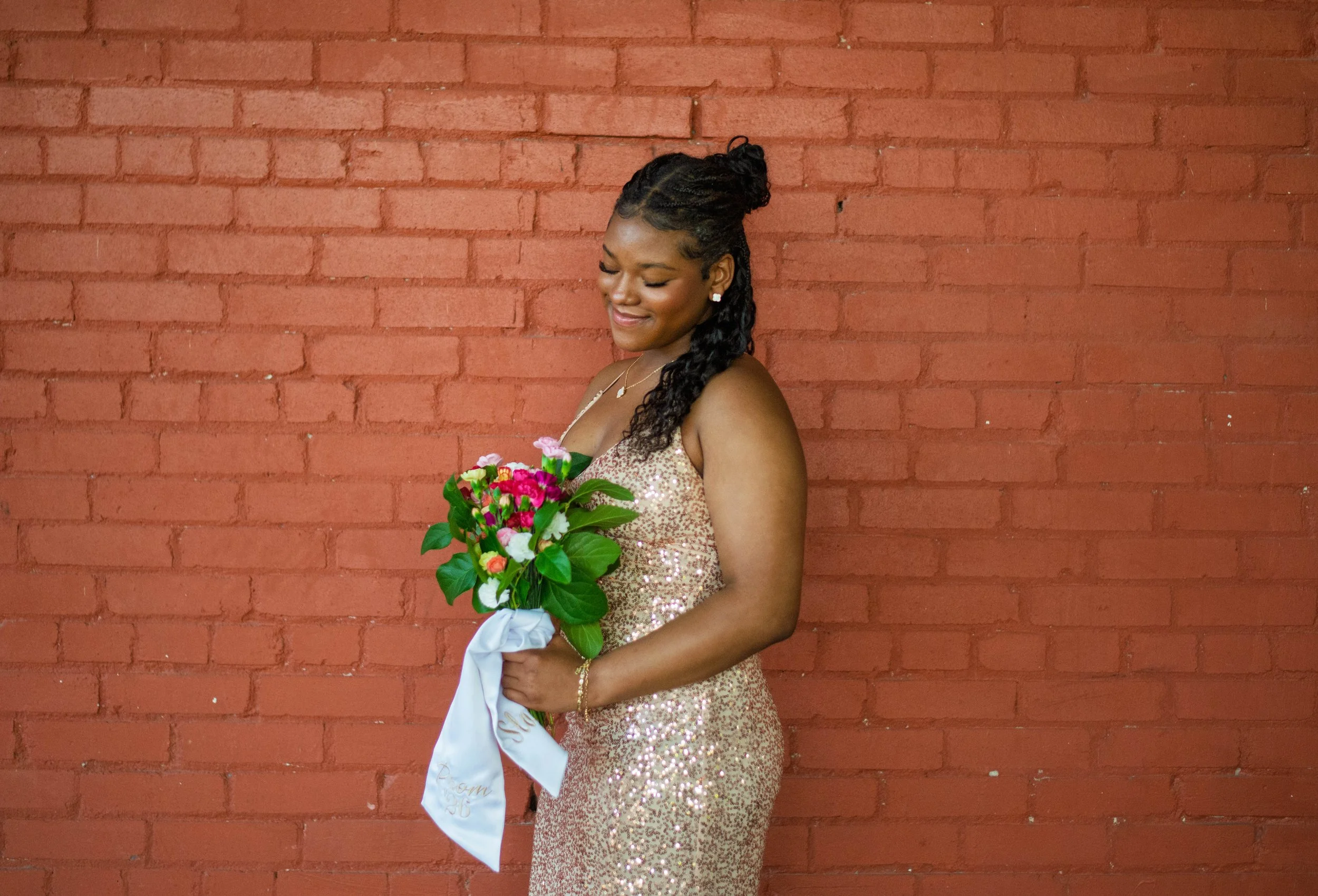 A woman in a sparkly, gold, sleeveless dress stands against a red brick wall, holding a colorful bouquet of flowers wrapped in white satin. She is smiling with her eyes closed.