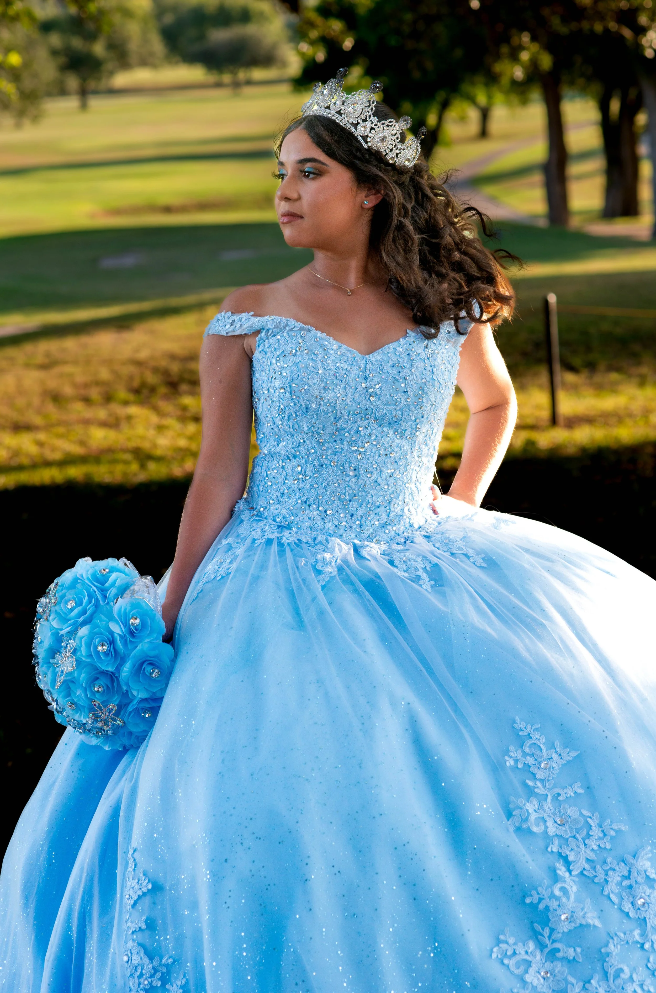 Young woman dressed as a princess in a light blue gown with lace and glitter, wearing a silver crown, holding a matching blue rose bouquet, outdoors in a park with trees and greenery.