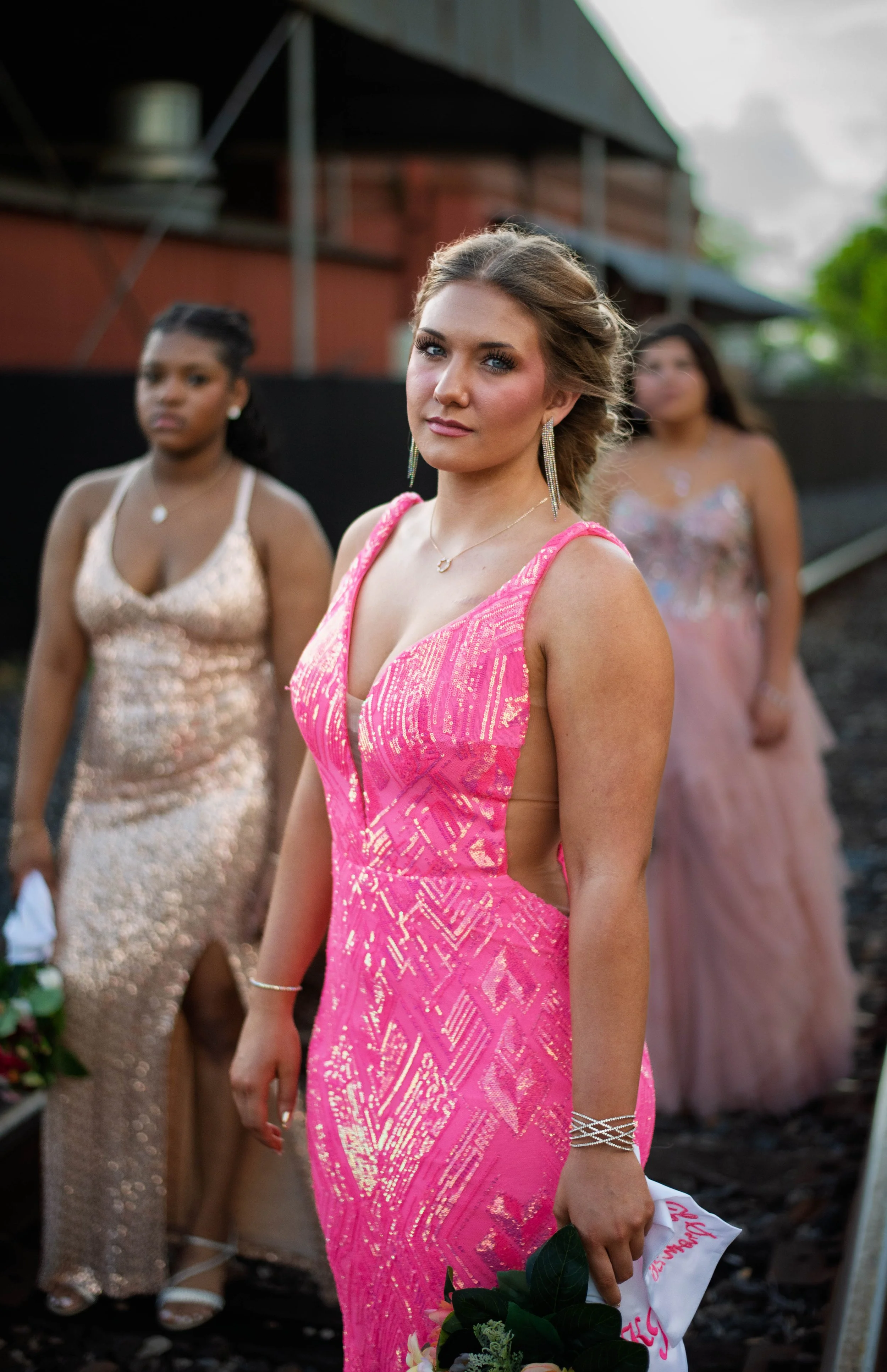 A young woman in a bright pink evening gown stands in the foreground, holding a bouquet of flowers, with two women in elegant dresses behind her outdoors.