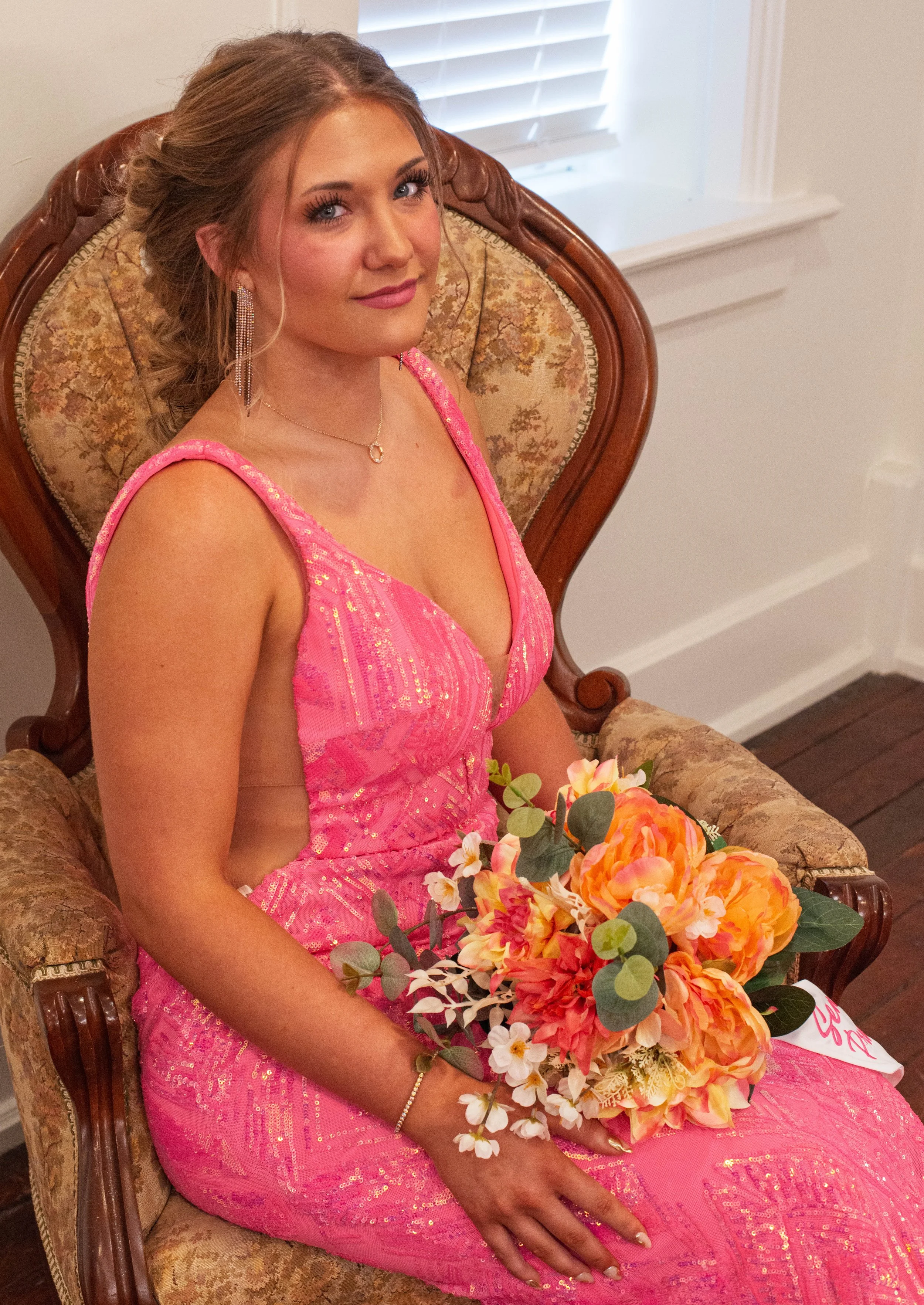 A young woman in a pink dress sitting on a vintage armchair holding a bouquet of flowers.