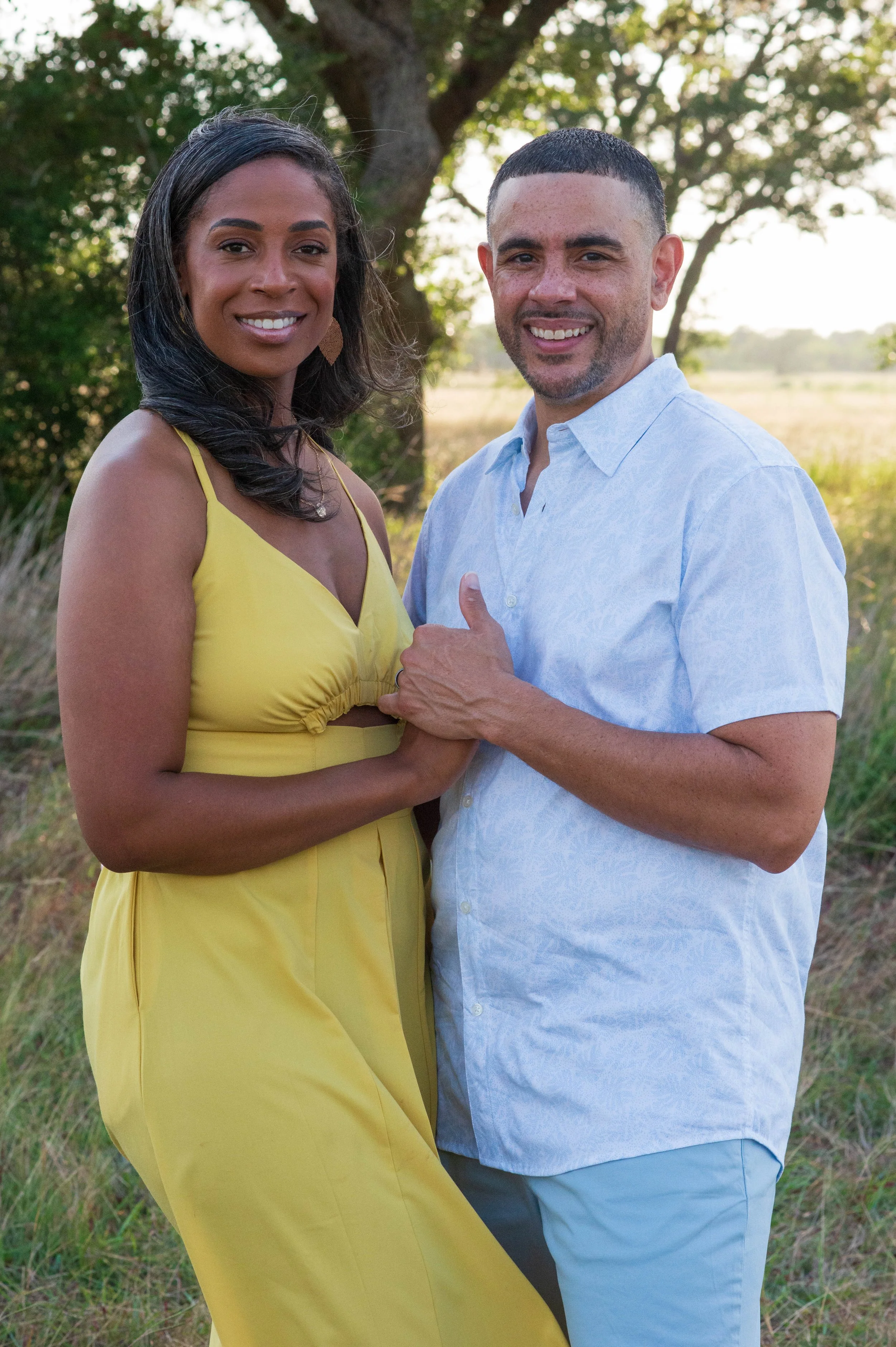 A smiling couple holding hands outdoors on a sunny day with trees and open field in the background.