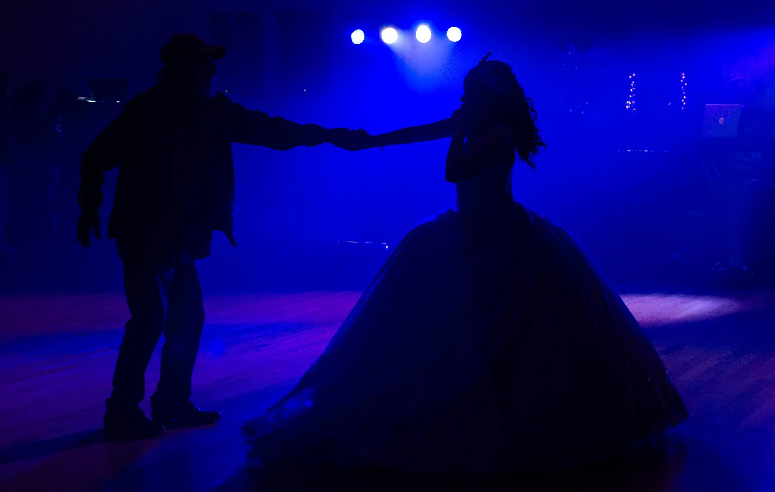 Silhouettes of a man and woman dancing, the woman in a large dress, in a dark room illuminated by blue lights.