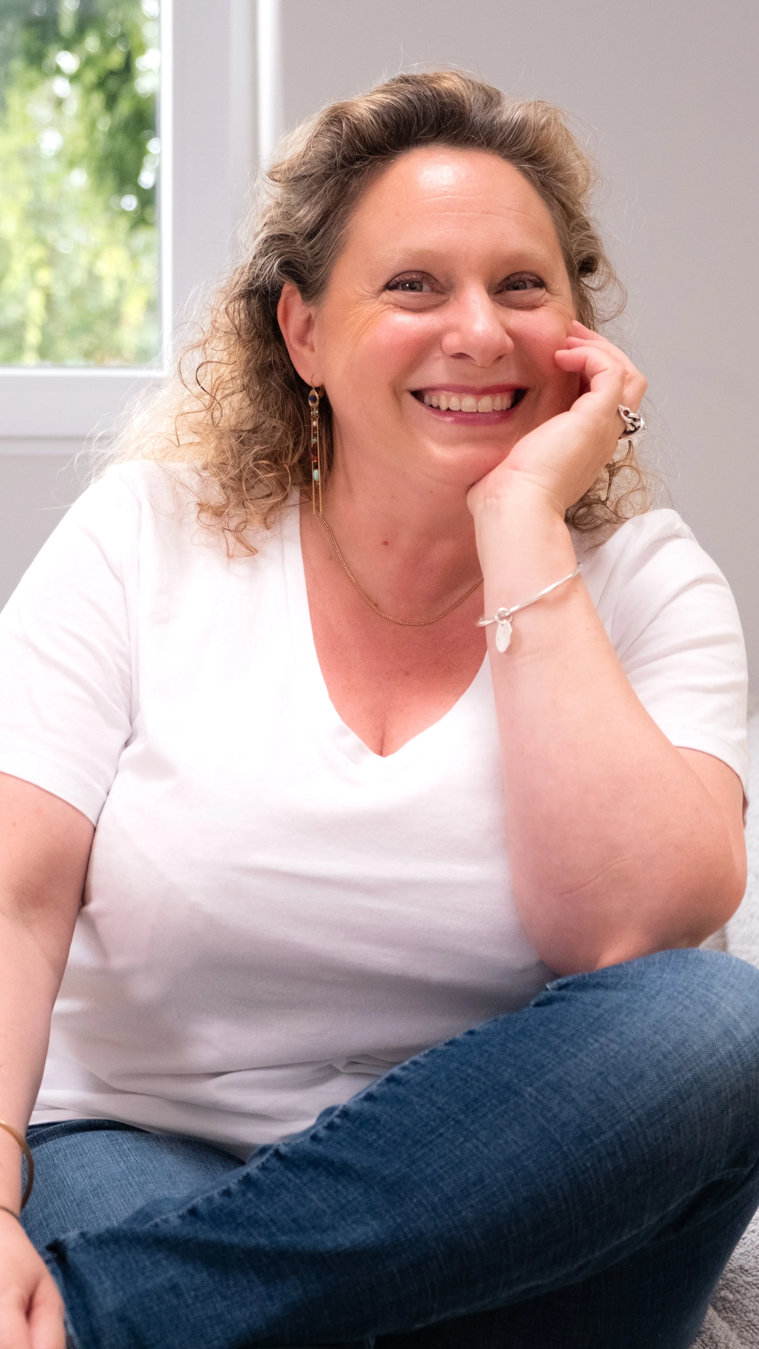 A woman with curly hair smiling and sitting cross-legged in front of a window with green foliage outside.