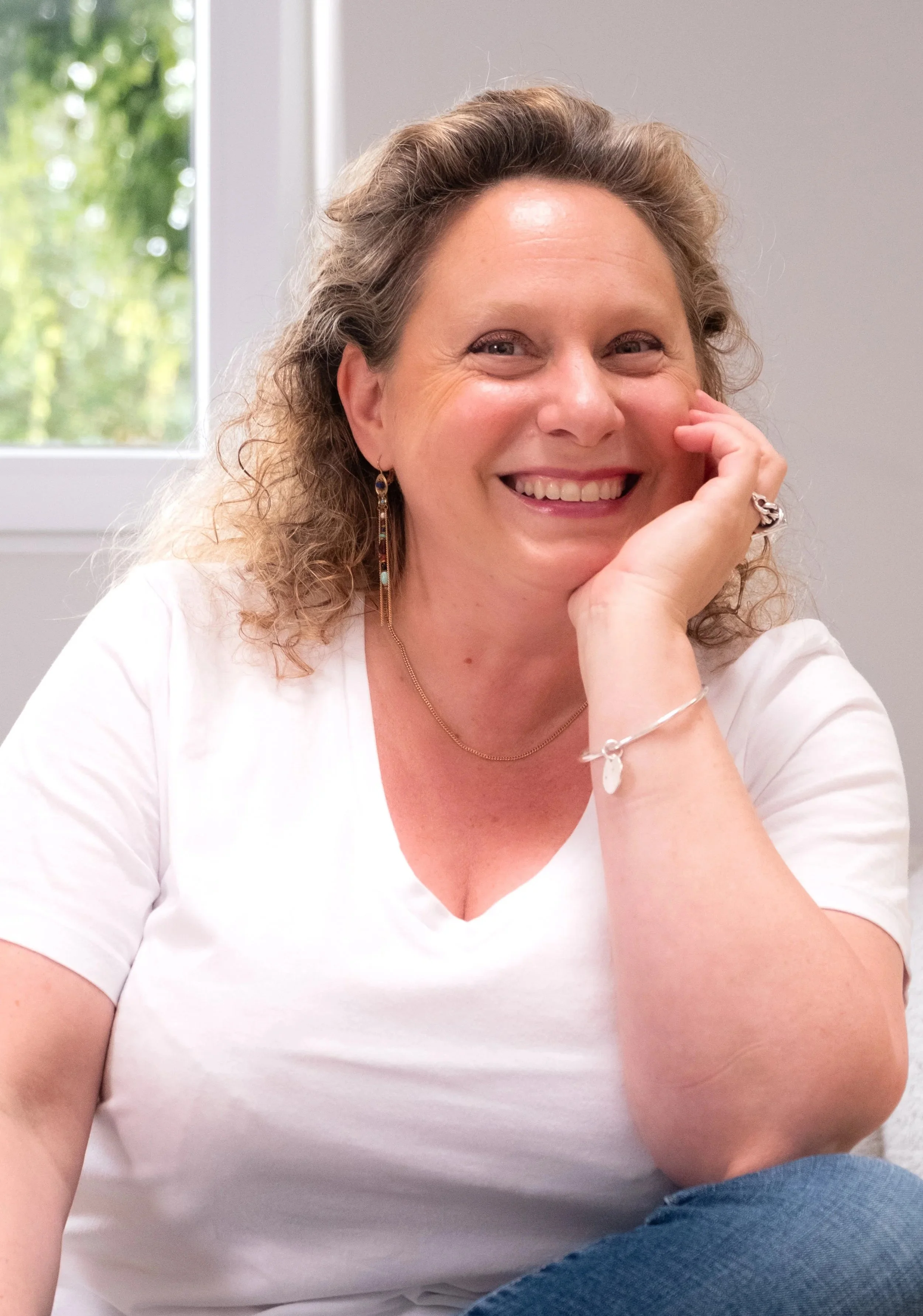 Smiling woman with curly hair wearing a white shirt, earrings, and a bracelet, sitting near a window with greenery outside.