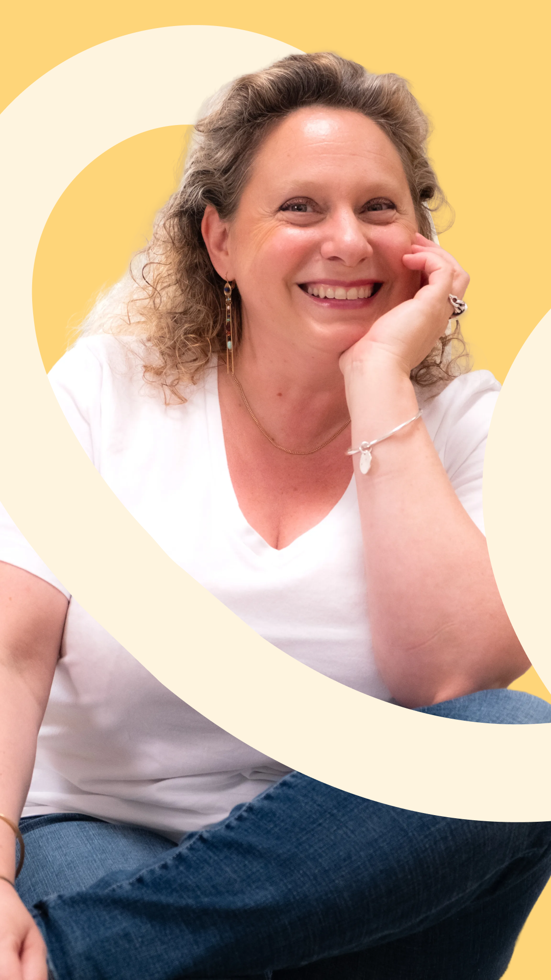 A smiling woman with curly gray hair, wearing a white T-shirt, blue jeans, and jewelry, sitting with her hand resting on her cheek, against a yellow background with white abstract shapes.