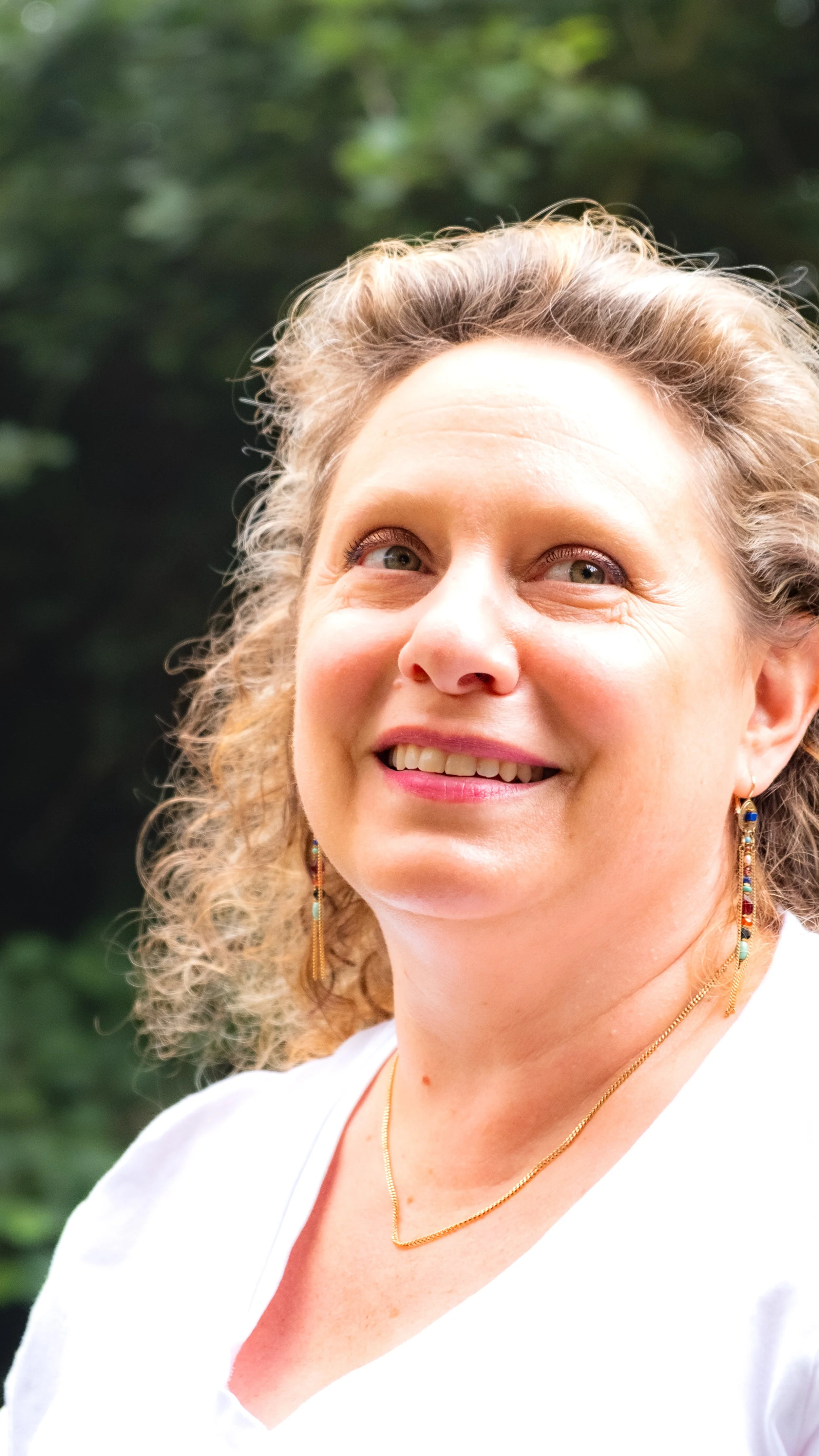 Close-up of a smiling woman with curly blonde hair, wearing earrings and a gold necklace, outdoors with green foliage in the background.