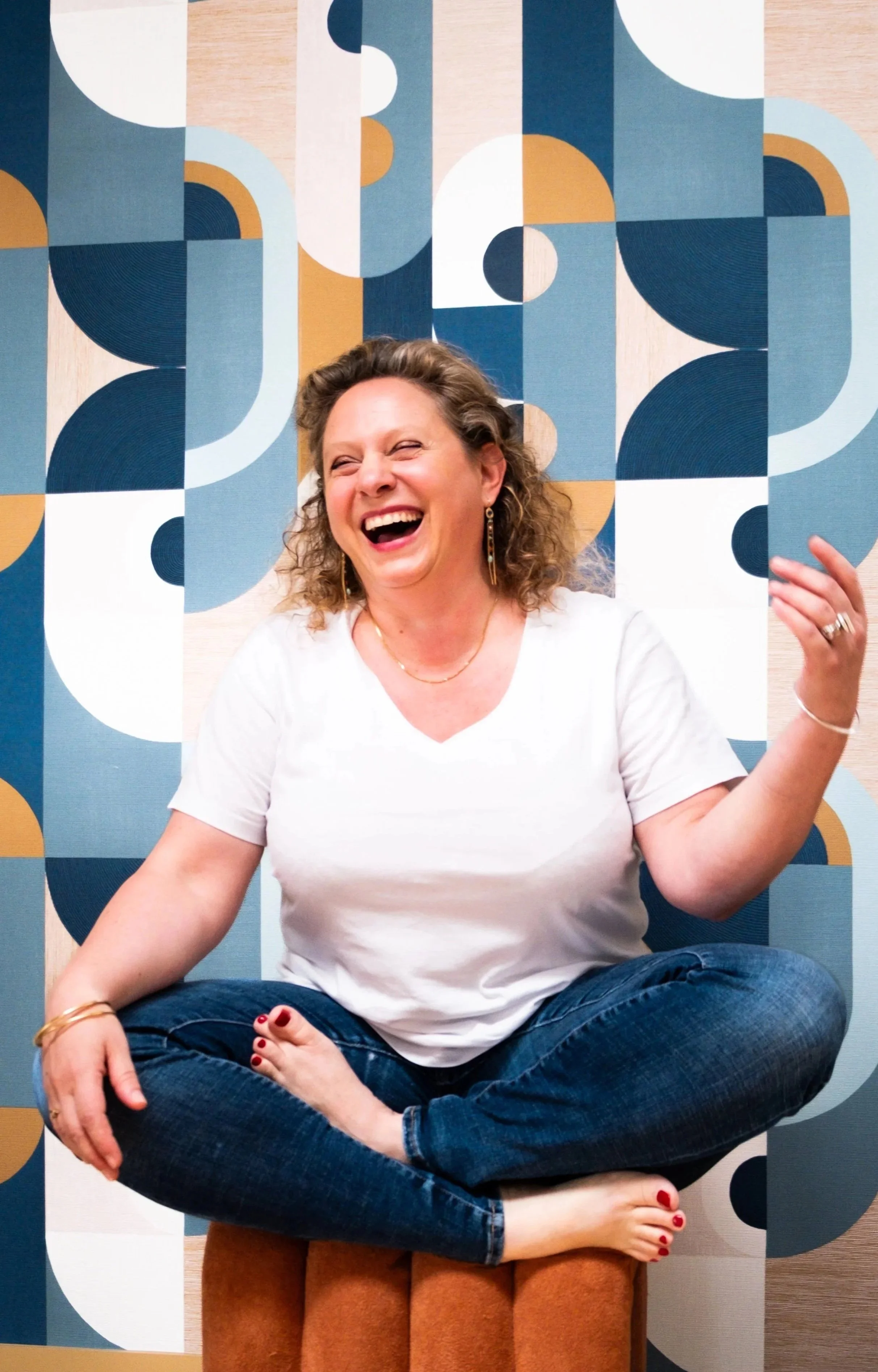 A woman sitting cross-legged on a stool, smiling and laughing, with a colorful abstract geometric mural in the background.