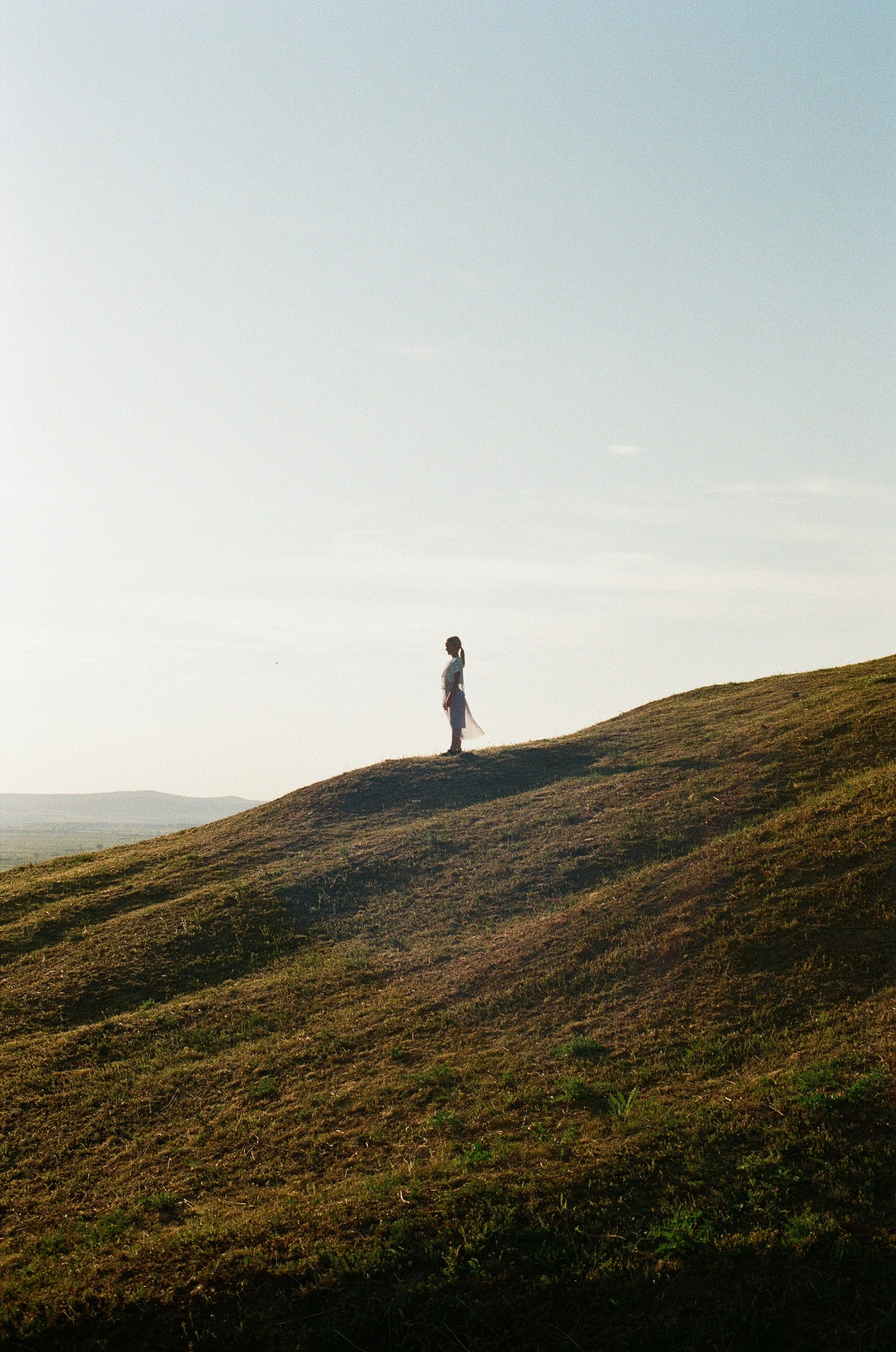 A person standing on a grassy hill with a view of distant mountains and a clear sky in the background.