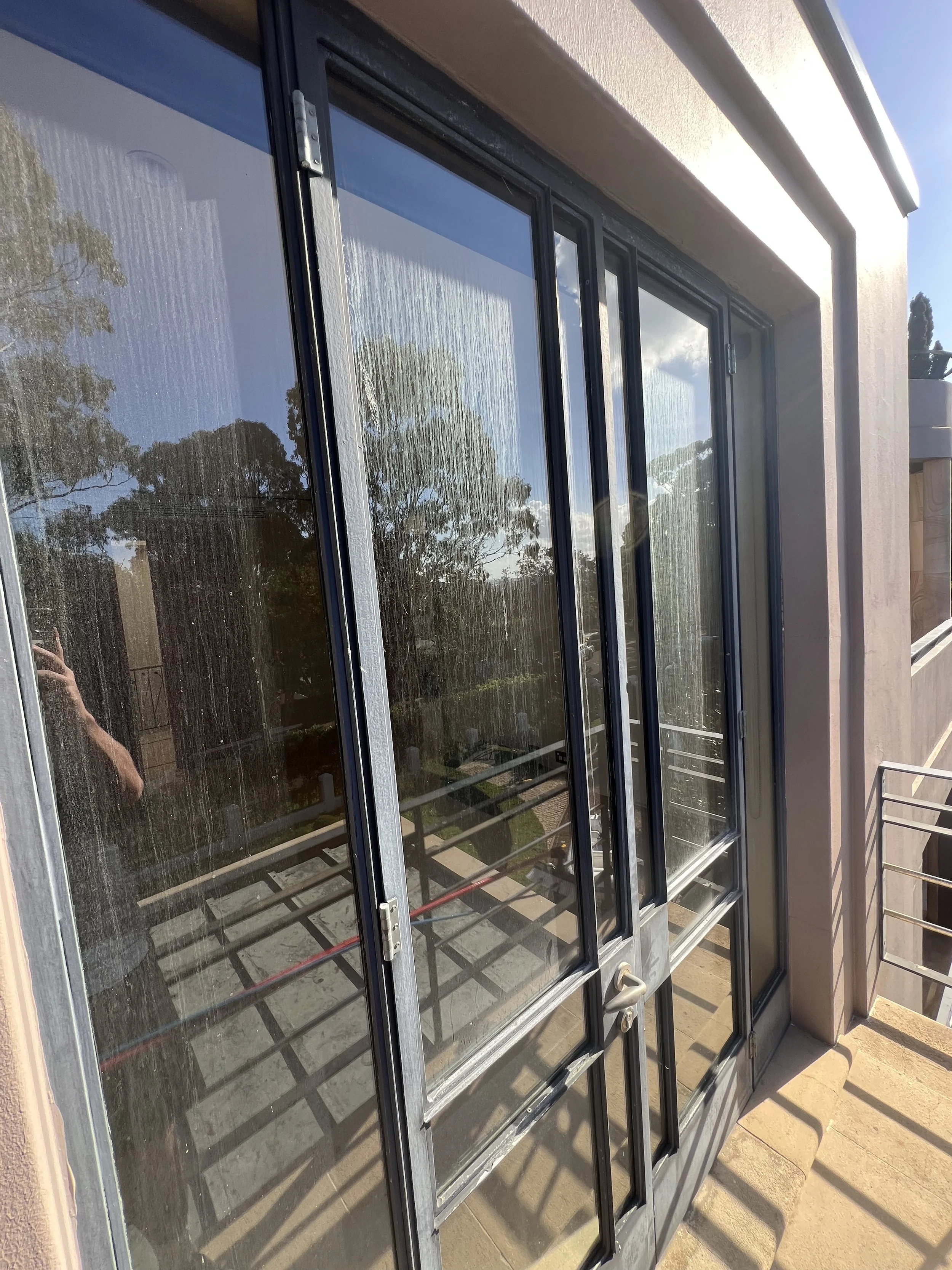 A sliding glass door with a black frame and handle, with glass reflecting trees, sky, and clouds, leading to an outdoor area with a concrete pathway and a building wall.