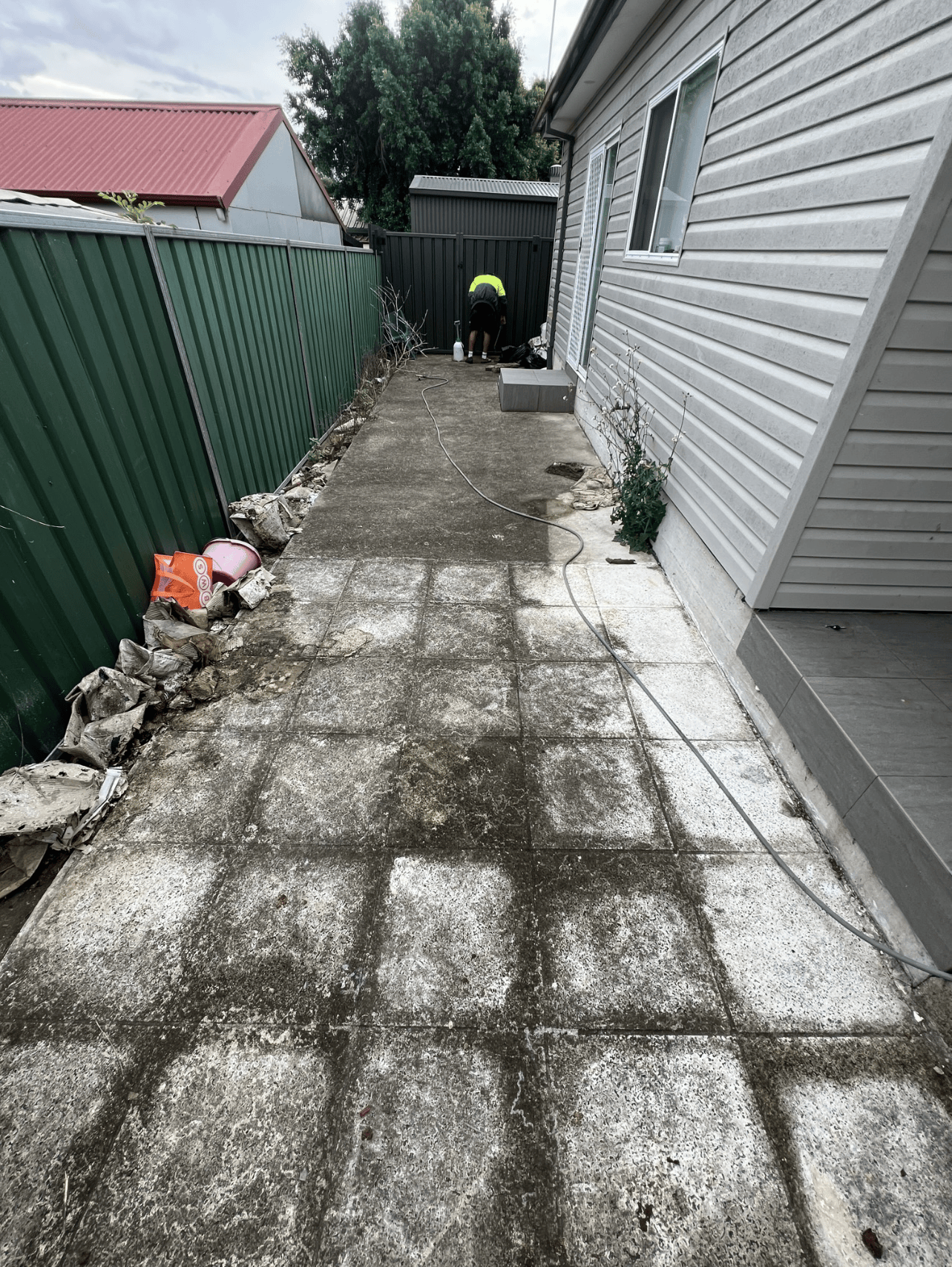 Backyard patio with concrete slabs, a person working near a gate, and a green and gray fence.