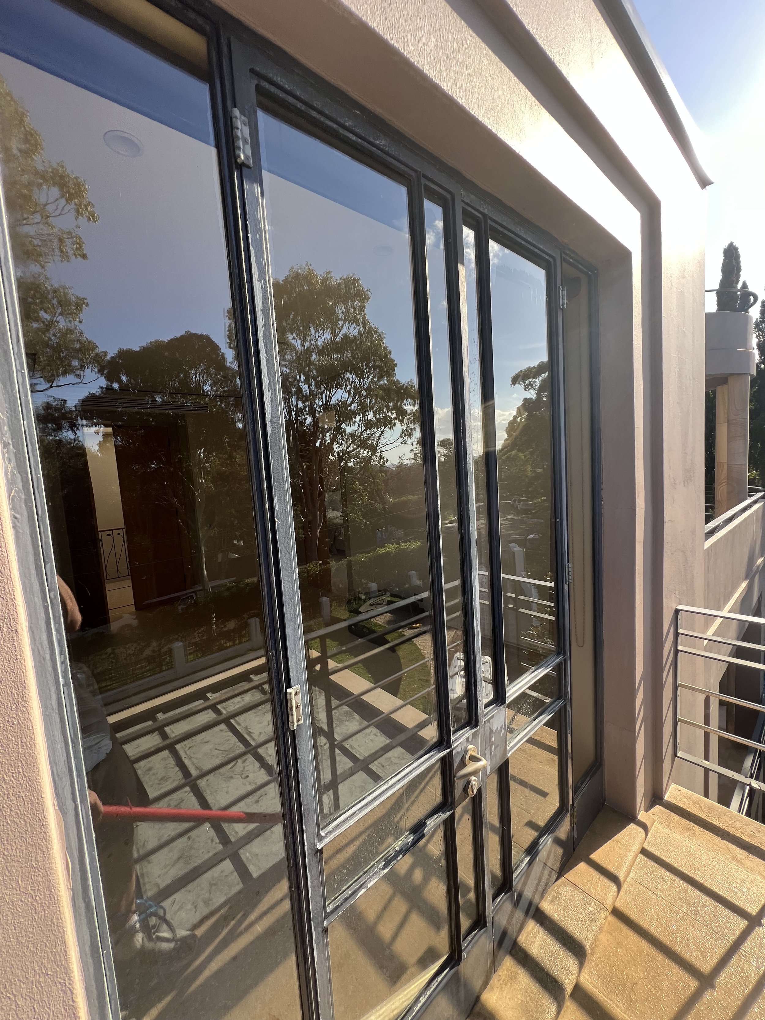 Close-up of a glass door with a black metal frame, part of a balcony or porch, with trees, sky, and clouds reflecting on the glass, and sunlight casting shadows on the ground.