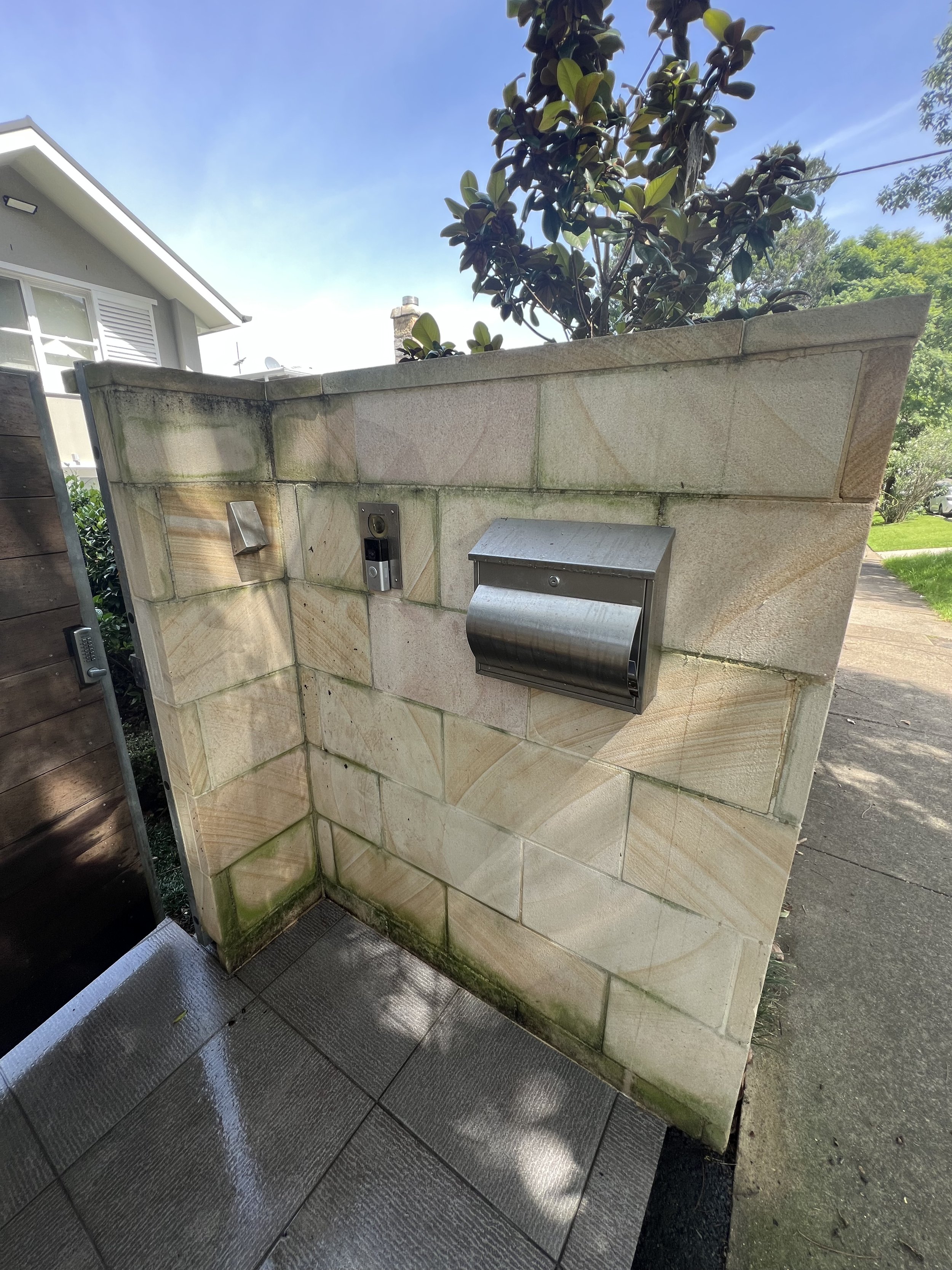 A stone wall with a mailbox, doorbell, and intercom, next to a sidewalk and house, with a building and green trees in the background under a blue sky.