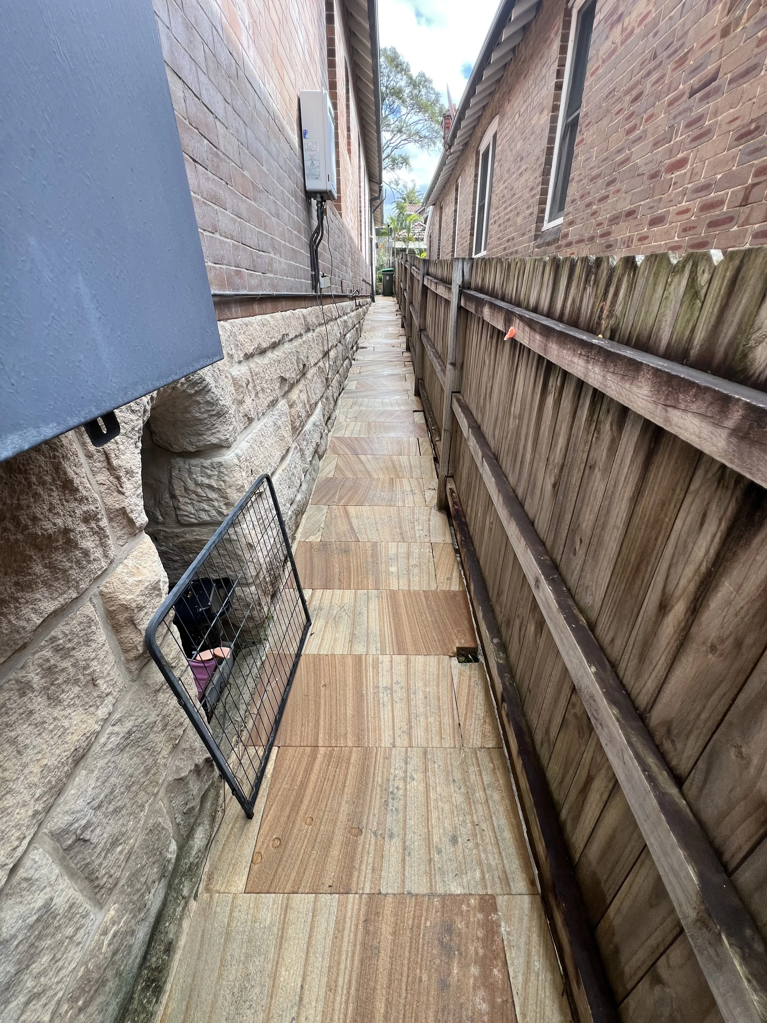 Side alley between brick houses, tiled concrete pathway, wooden fence on the right, utility box on the left, some trees and a cloudy sky in the background.