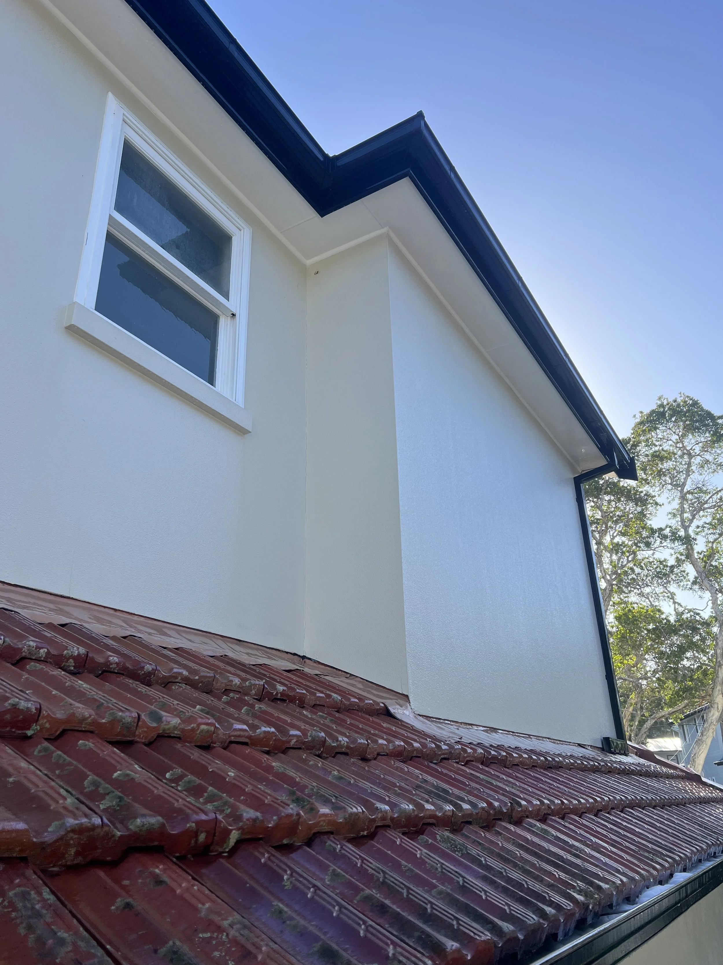 Close-up of a house's white exterior wall with a small rectangular window, a dark overflowing gutter, and a red tiled roof at the bottom of the image, with some trees and blue sky in the background.