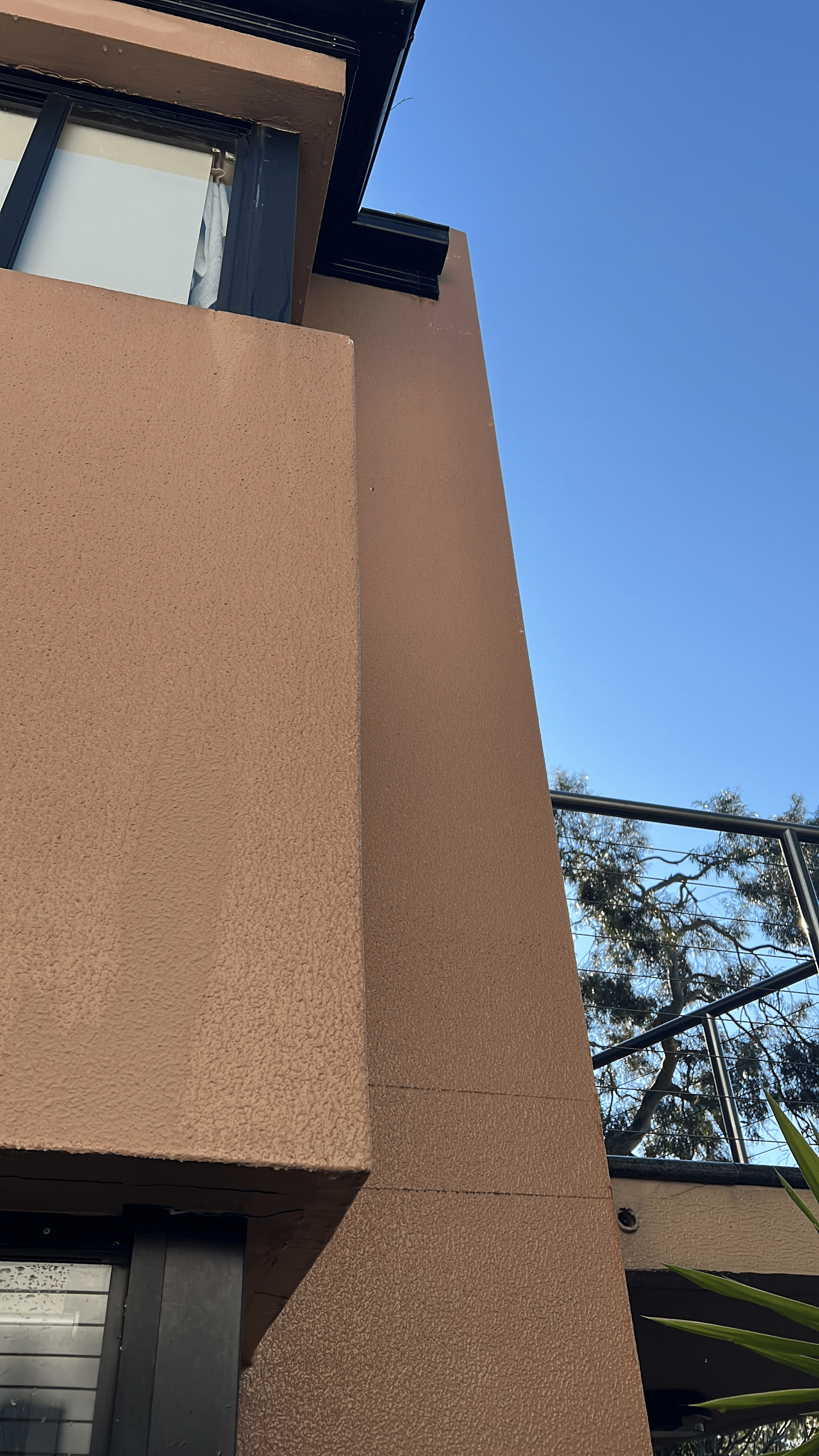 Close-up view of beige stucco exterior wall of a building with black trim around a window and a balcony railing against a bright blue sky.