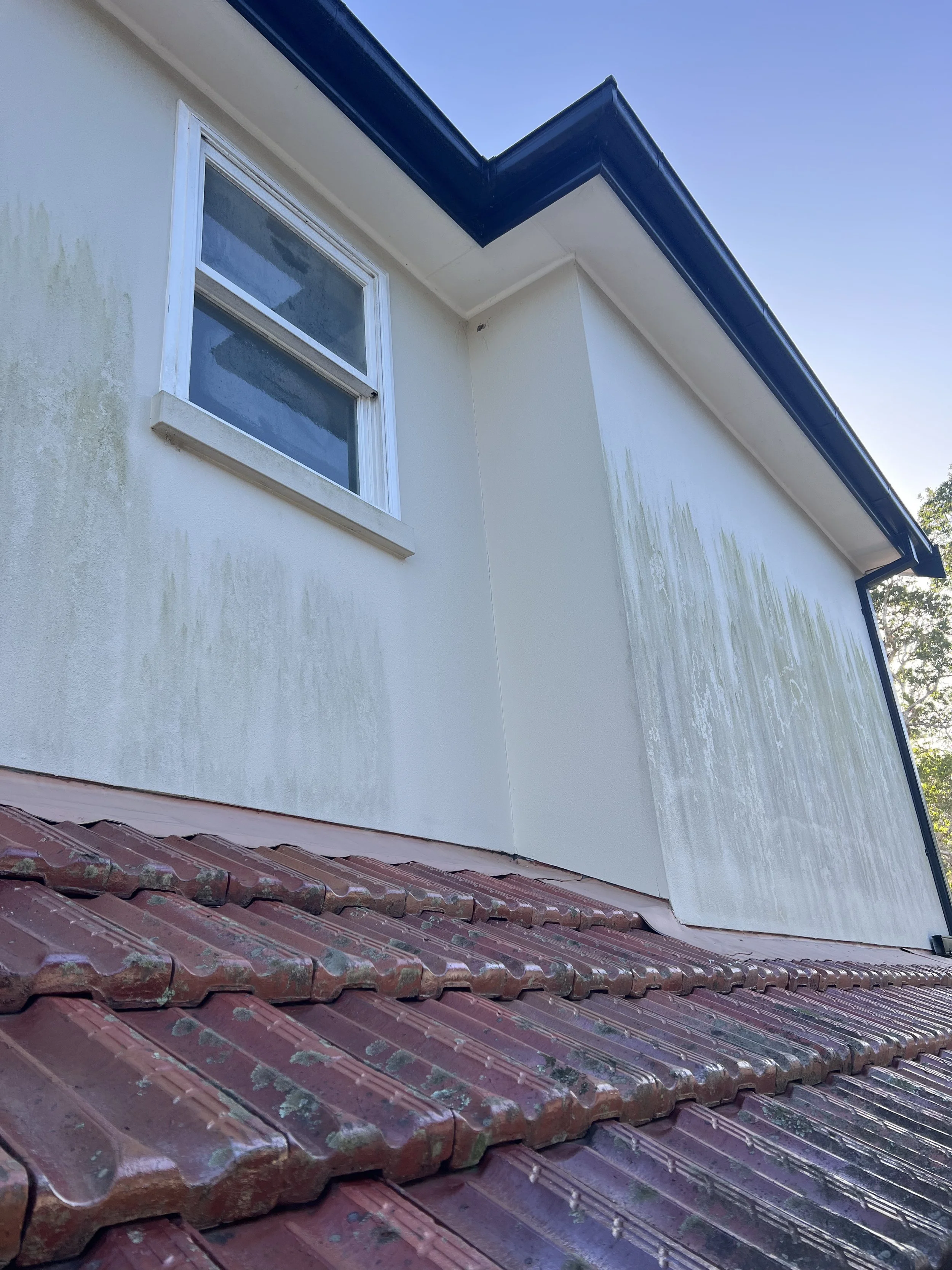 The image shows part of a house exterior with a window on a cream-colored wall. The house has a red-tiled roof, and the roofline features blue trim and white soffits under the eaves. The wall has some visible weathering or staining below the window.