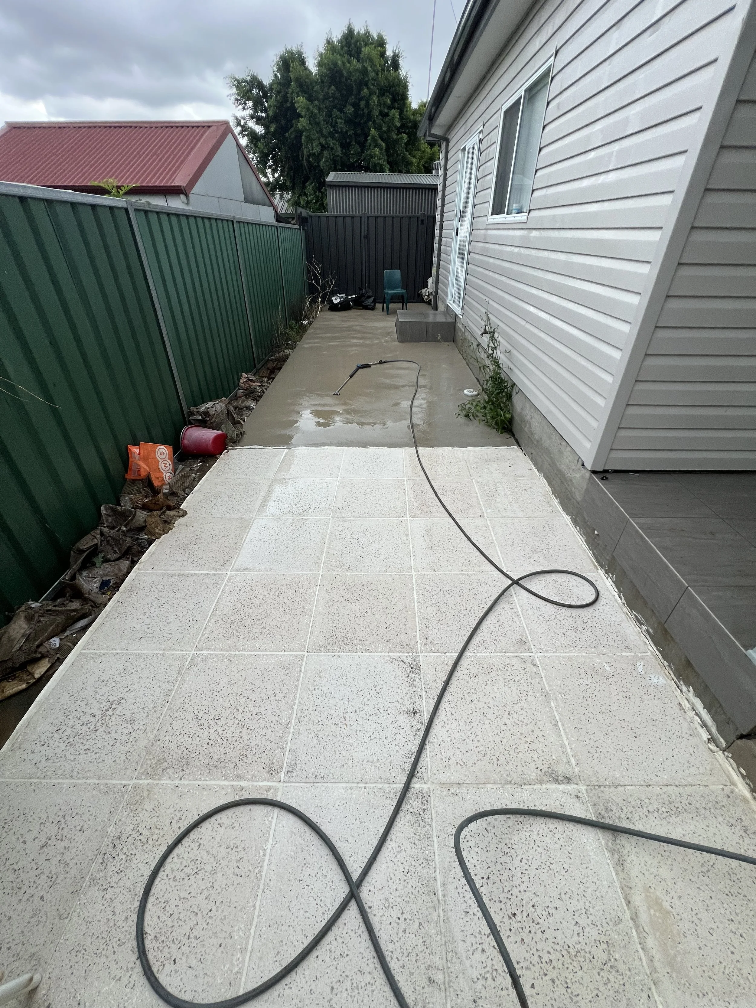 The image shows a backyard patio with a concrete floor, partially covered with wet tiles, adjacent to a white house with vinyl siding. There is a black garden hose lying on the concrete, an orange bag and a red container on the left, and some garden 