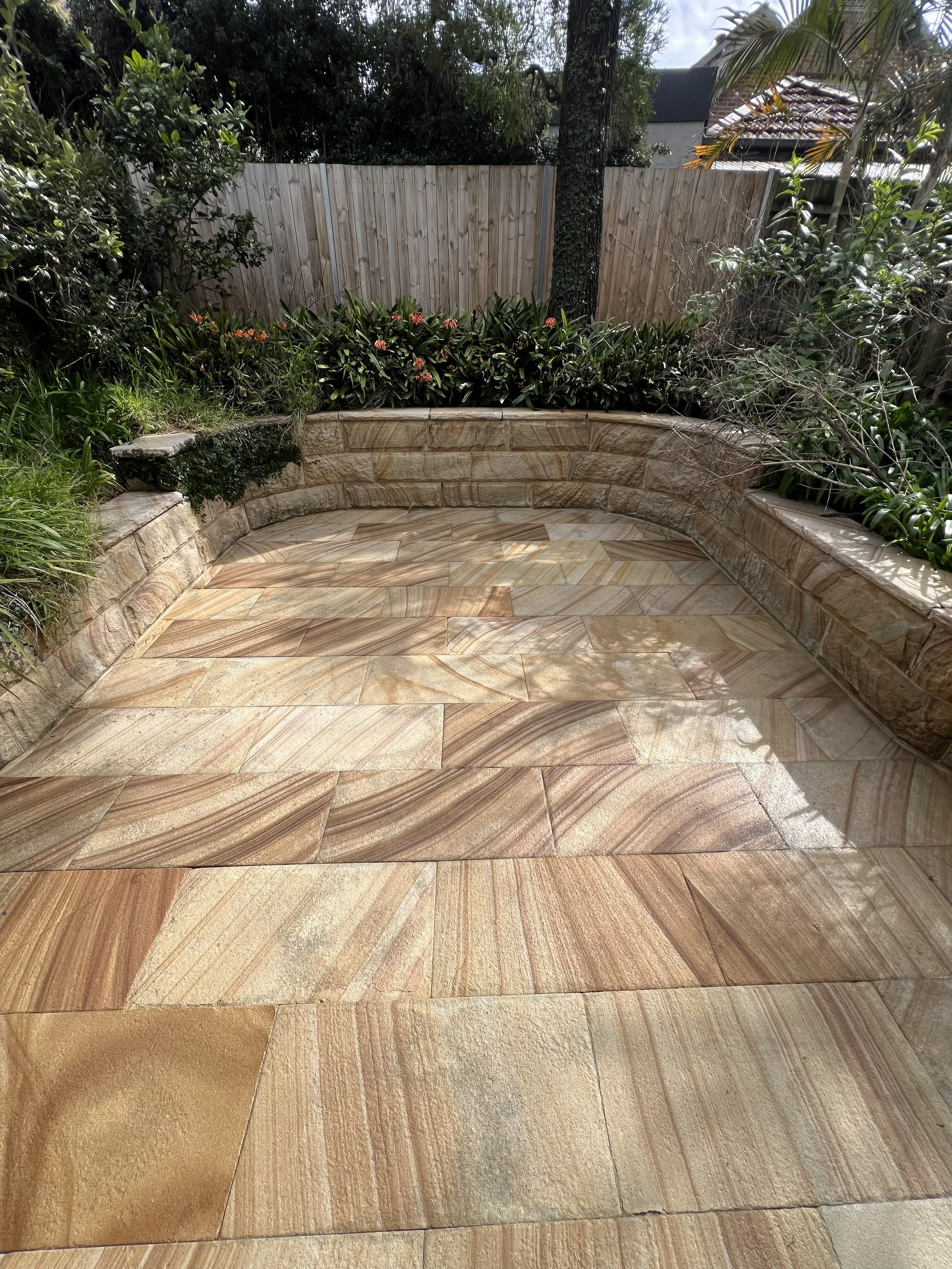 A backyard area with a curved stone bench and a tiled stone floor. There are plants and bushes along the back, a wooden fence, and some trees and a house in the background.
