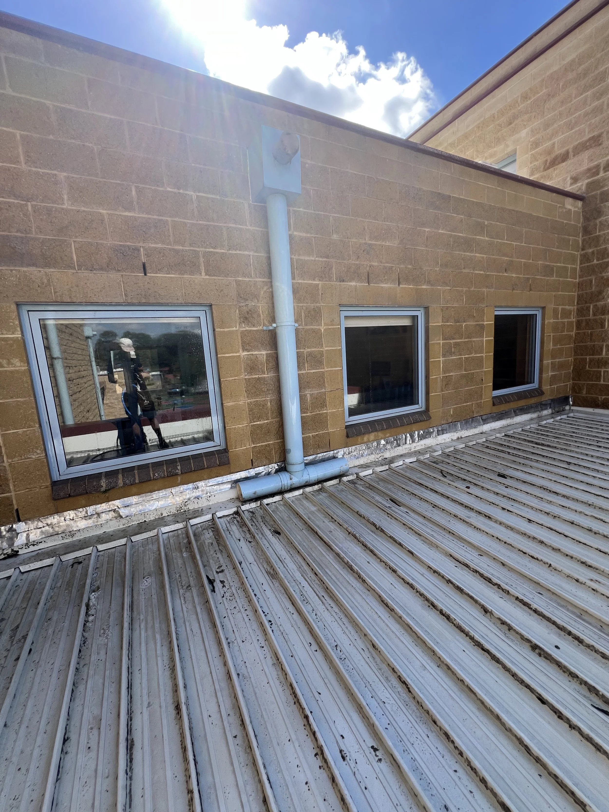 Exterior view of a brick building with three small windows and a metal pipe running along the wall. Reflection of a person taking the photo is visible in one window, sunlight is visible at the top of the image, and the sky is partly cloudy.