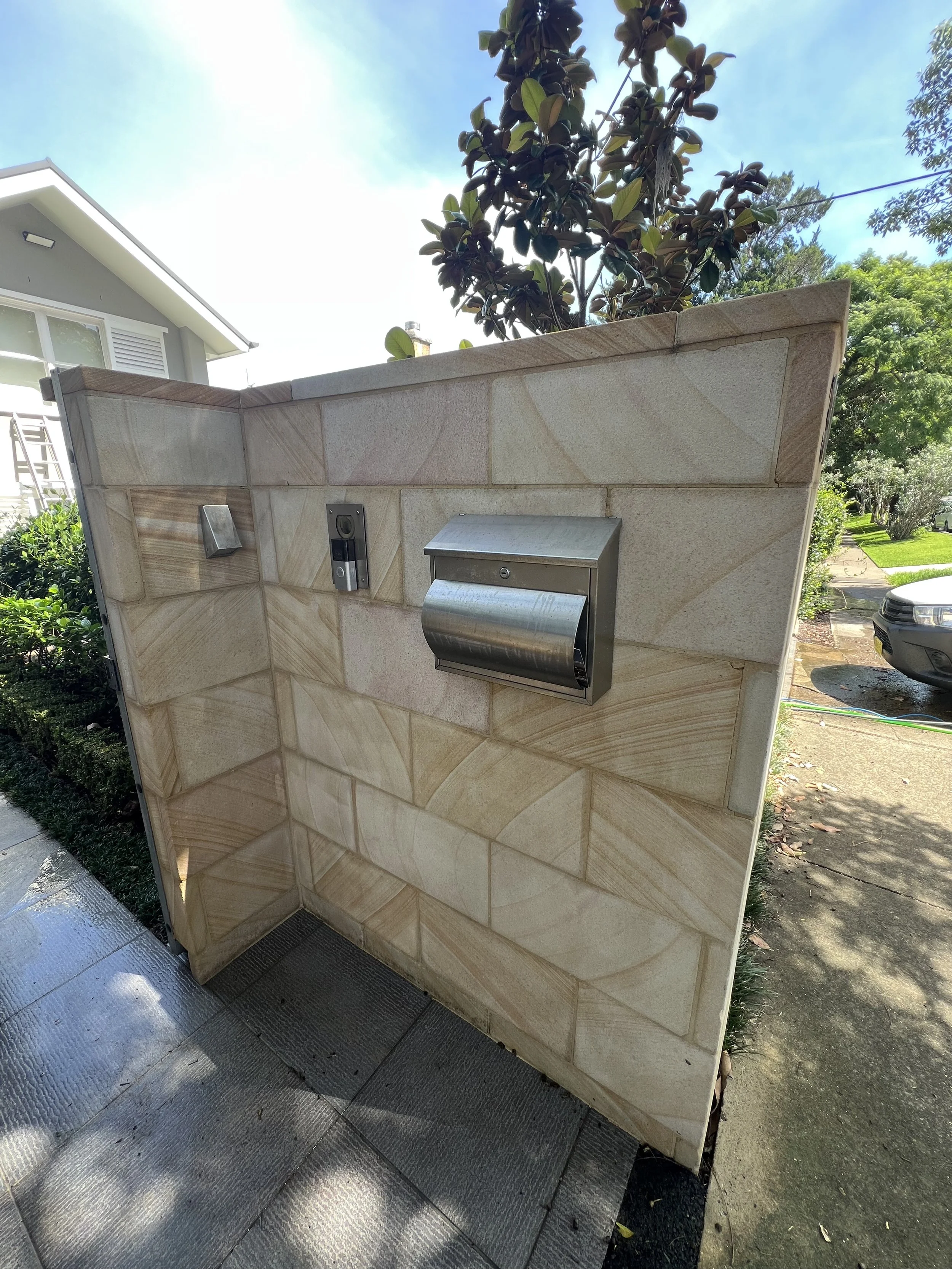 Outdoor view of a beige tiled wall with a mailbox and intercom, background of trees, a sidewalk,  lawn, and a house with a white roof.