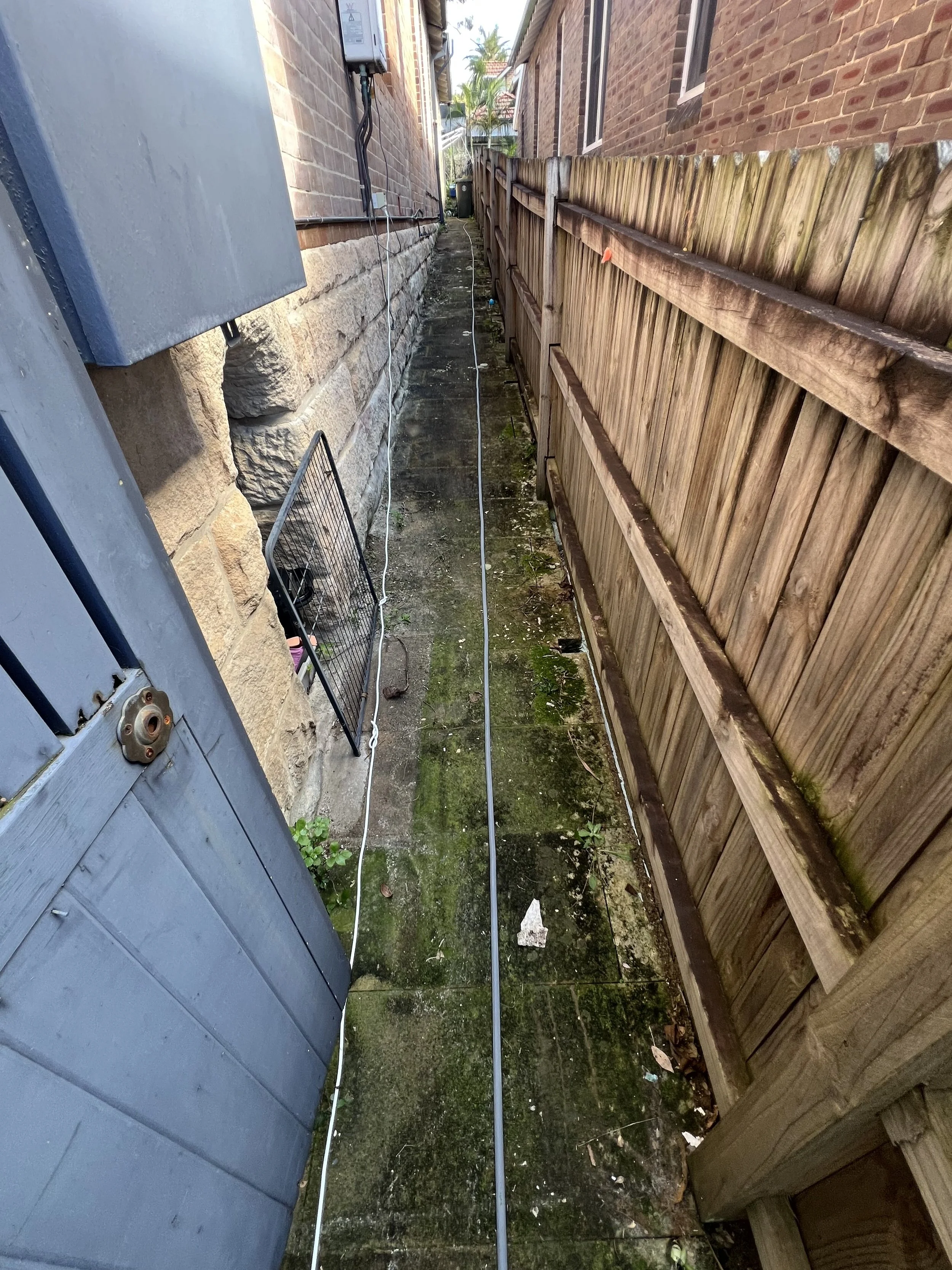 A narrow outdoor space between two brick buildings with a wooden fence on the right and a blue-grey utility box on the left. A power meter and electrical wiring are on the brick wall, and a black metal rack leans against the utility box. The ground i