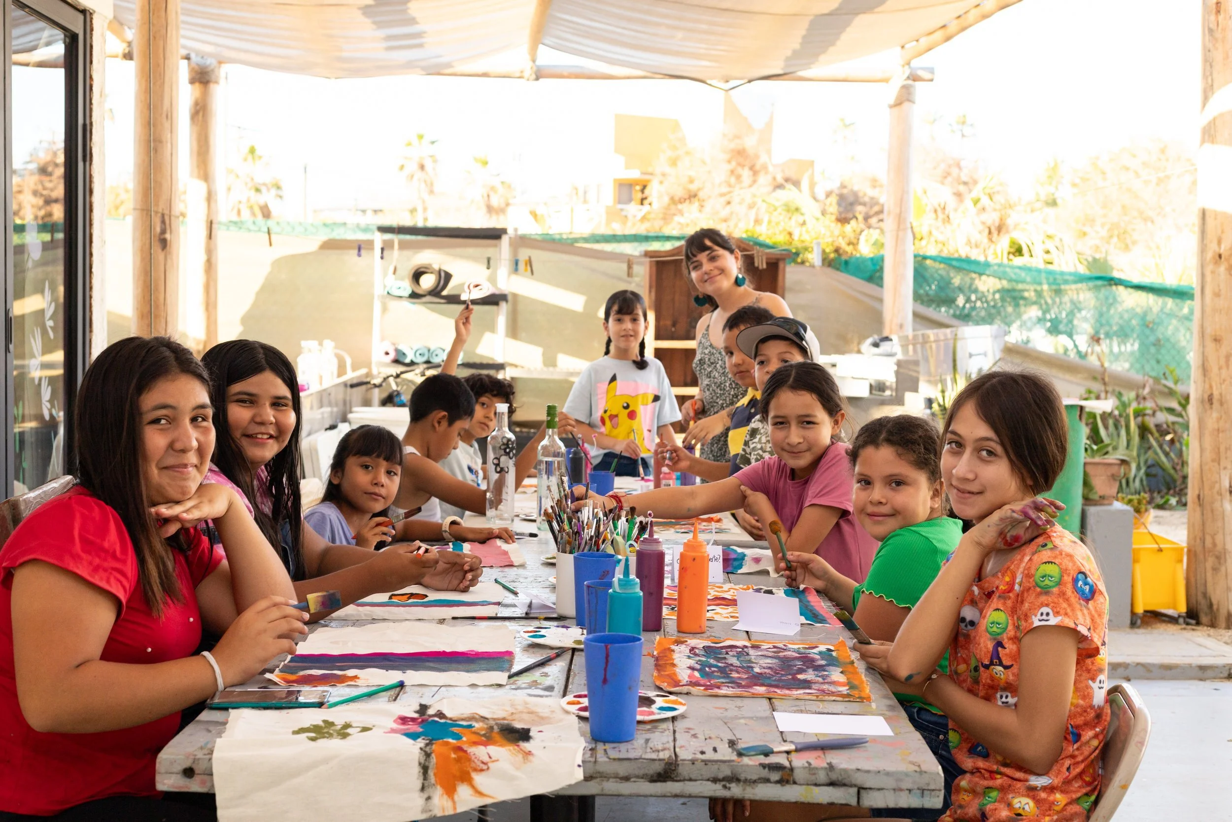 Niños y una mujer haciendo arte en una mesa larga en un espacio abierto, rodeados de materiales de pintura y decoraciones, en un ambiente alegre y soleado.