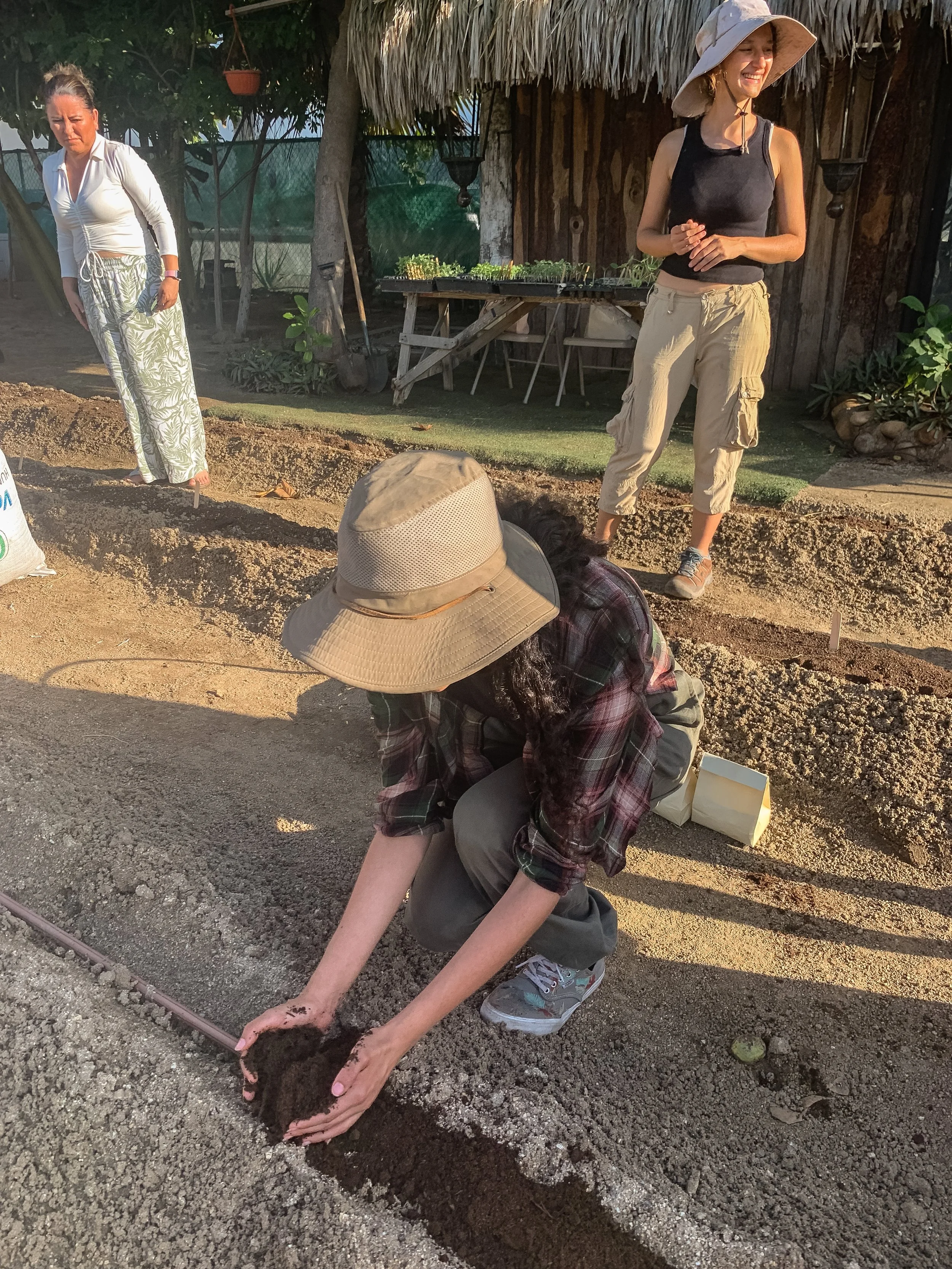 Mujer arrodillada en la tierra plantando con una mano en el suelo y la otra sosteniendo una herramienta de jardinería. Hay otra mujer de pie en el fondo. Escenario al aire libre con plantas, sombra y estructura de madera.