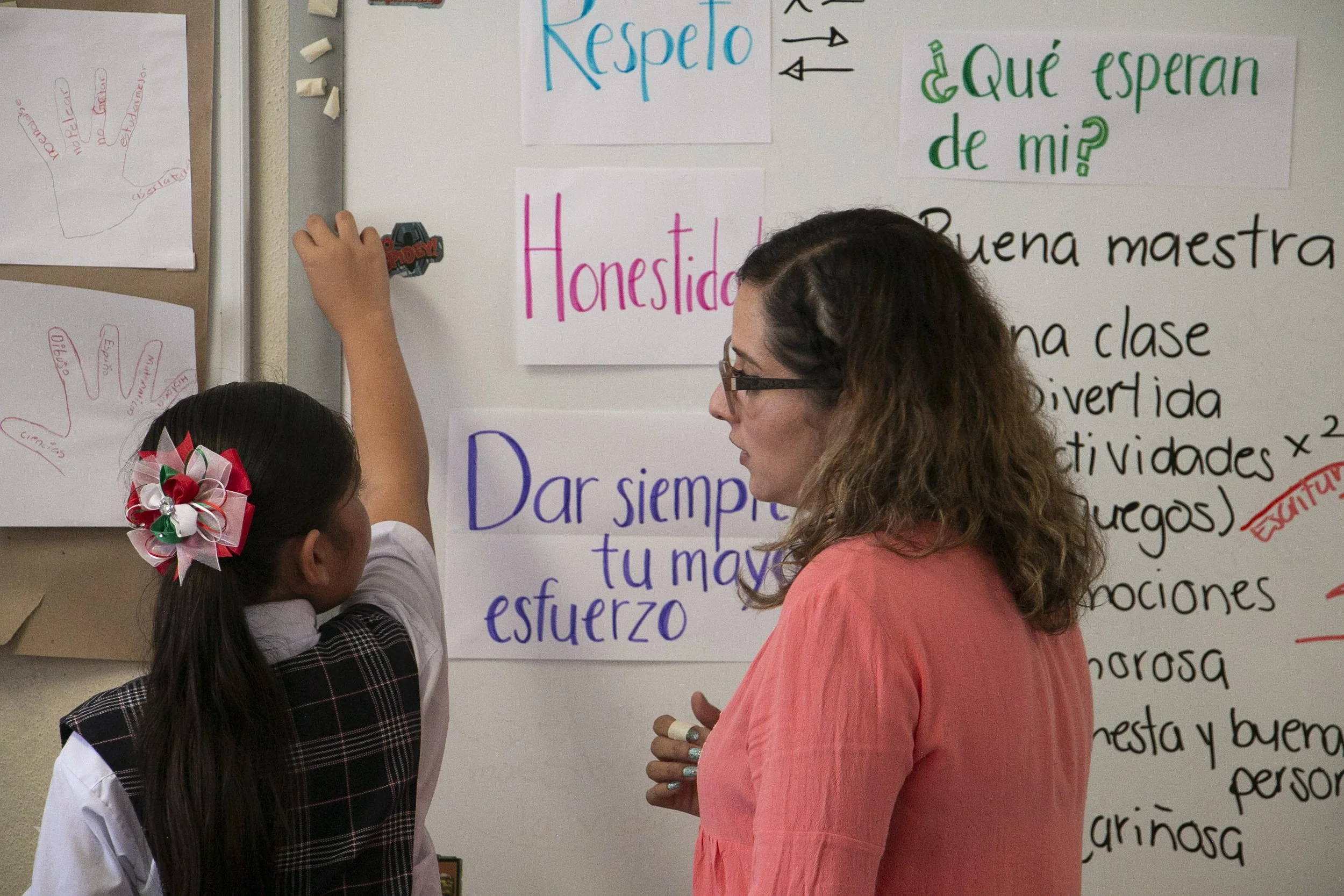 Una maestra y una niña hablando frente a una pizarra con carteles y escritura en letras de diferentes colores en un aula escolar.