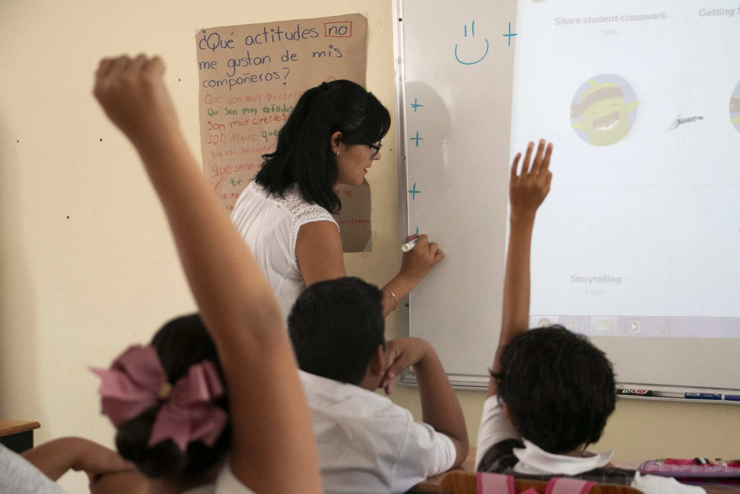 Una maestra enseña a una clase con estudiantes levantando la mano en un aula, en un pizarrón se puede leer una pregunta en español sobre actitudes hacia los compañeros.
